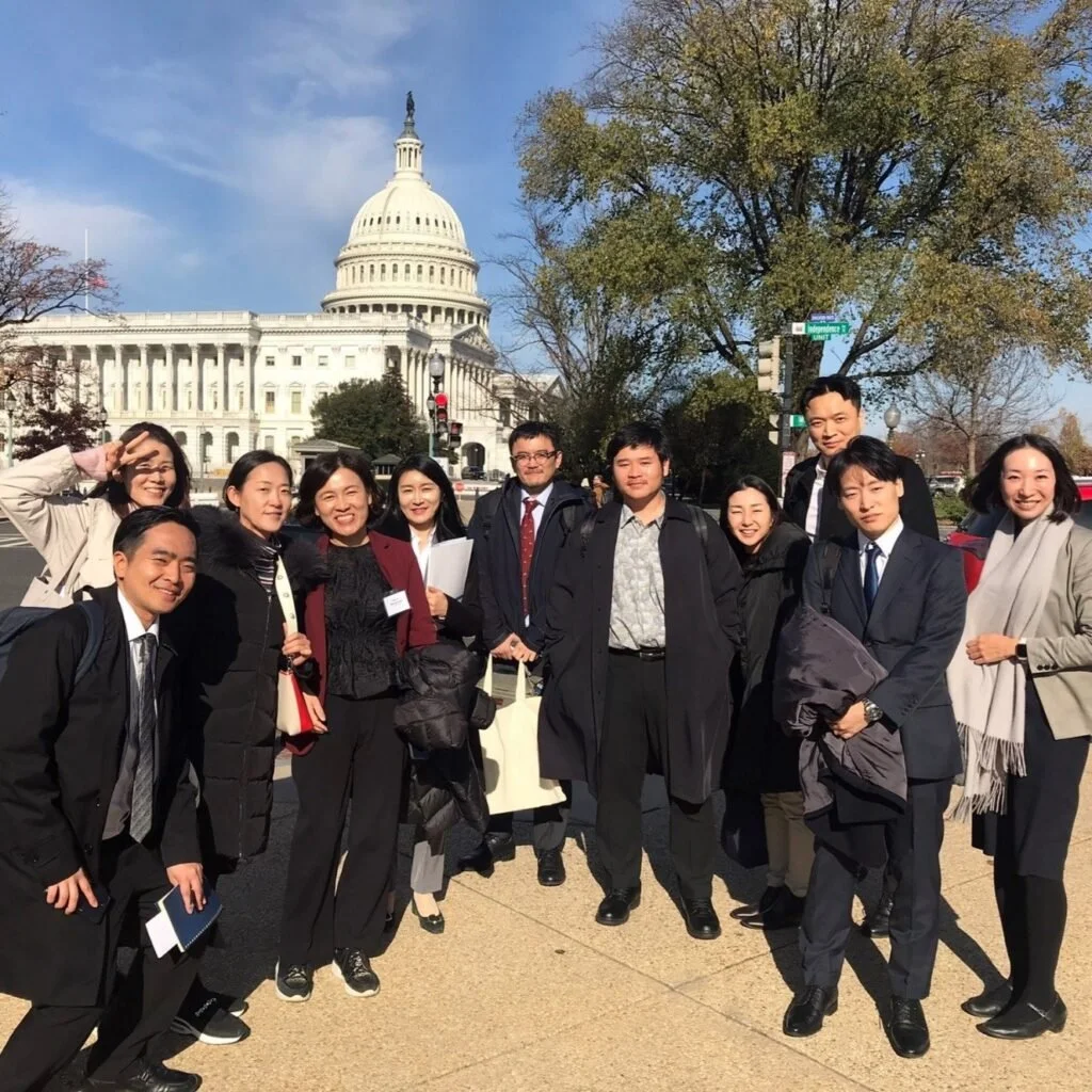 Group of diverse people standing in front of the U.S. Capitol building on a clear day with trees and a street sign visible.