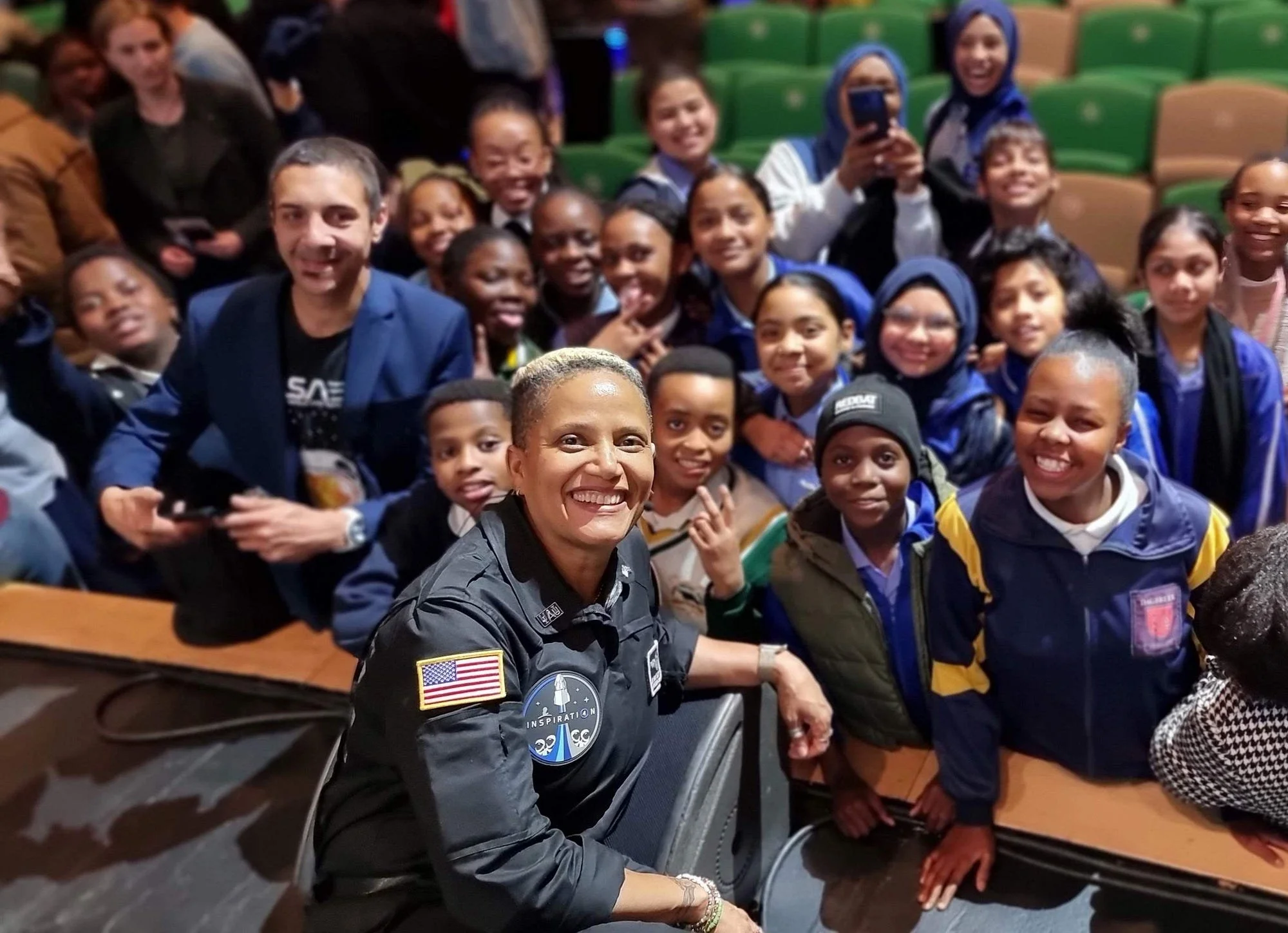 A group of young students smiling for a photo with a smiling woman in a black jacket in an auditorium or theater.