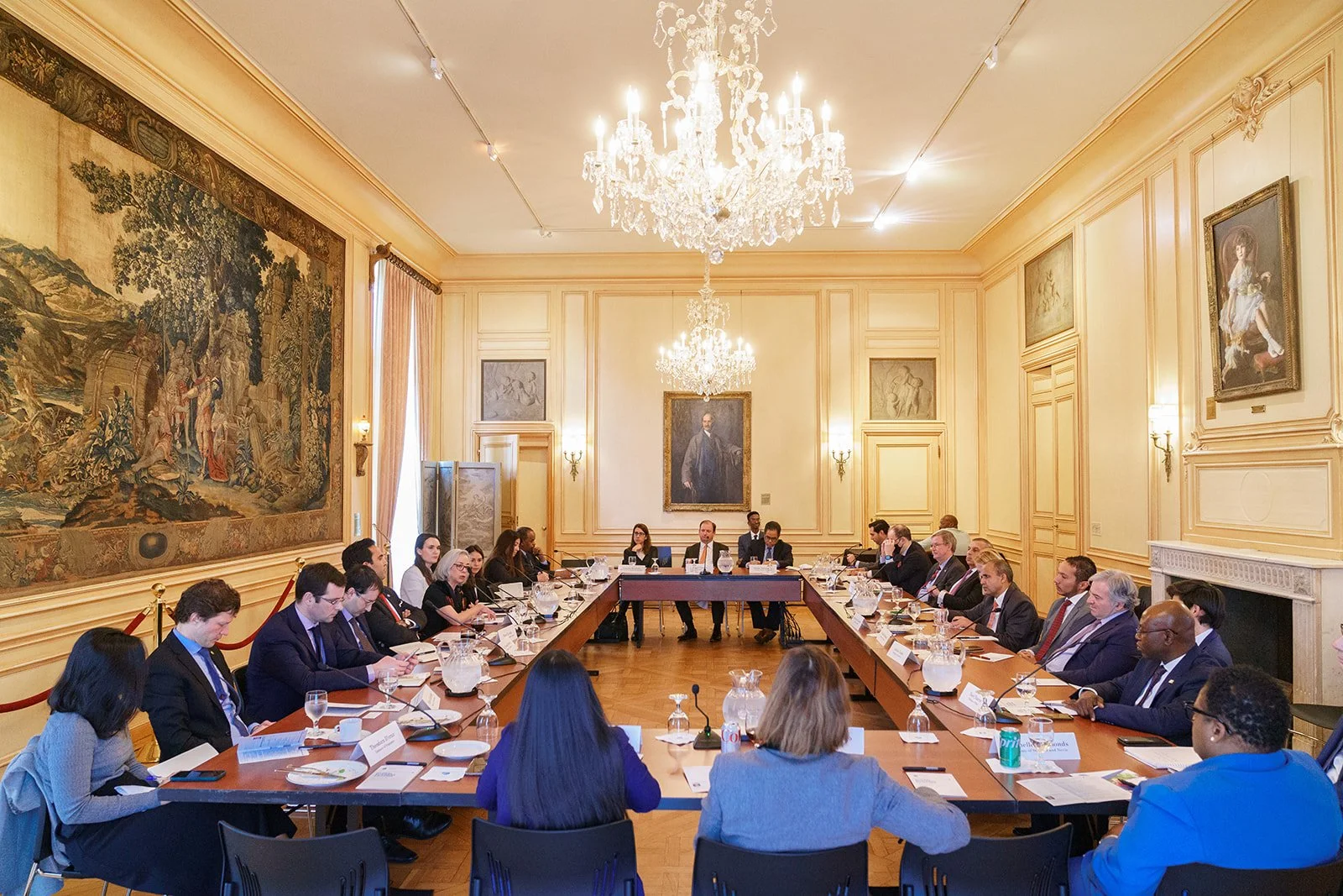 A formal meeting in an elegant, historical room with ornate walls, large paintings, chandeliers, and a long conference table surrounded by seated participants.