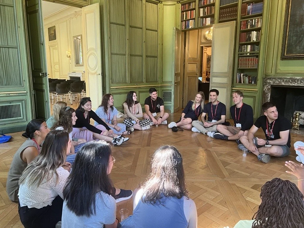 A group of young people sitting on the wooden floor in a circle inside a room with green paneled walls and built-in bookshelves.