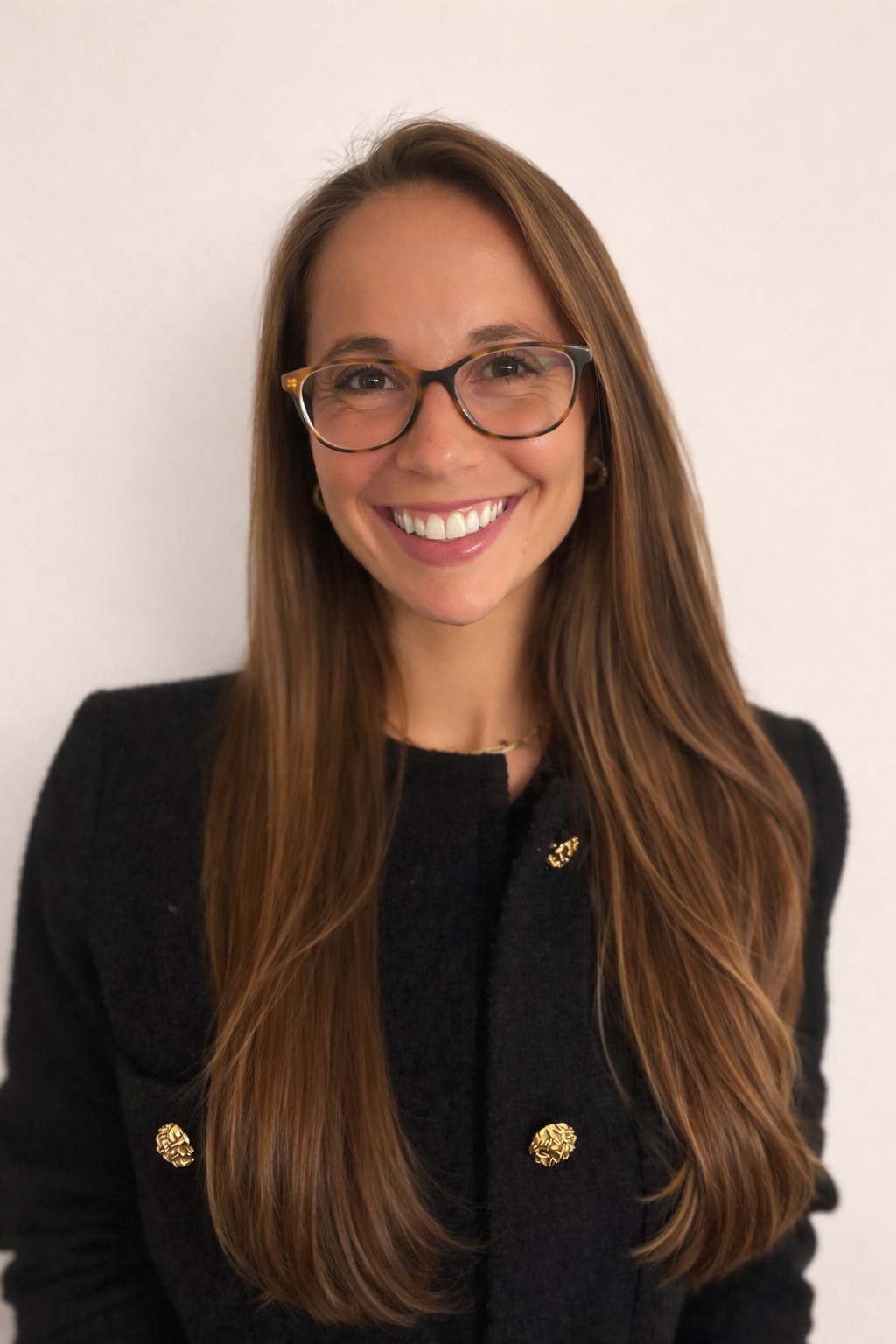 A smiling woman with long brown hair, glasses, and gold jewelry wearing a black blazer standing against a plain white wall.