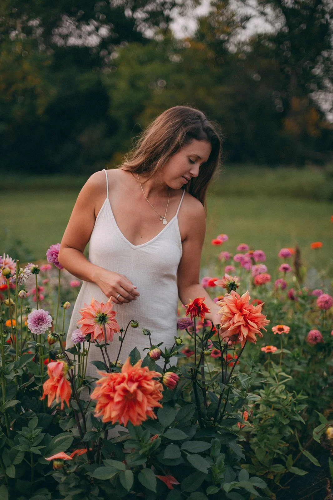 A woman with long brown hair in a white slip dress standing in a colorful flower garden during sunset.