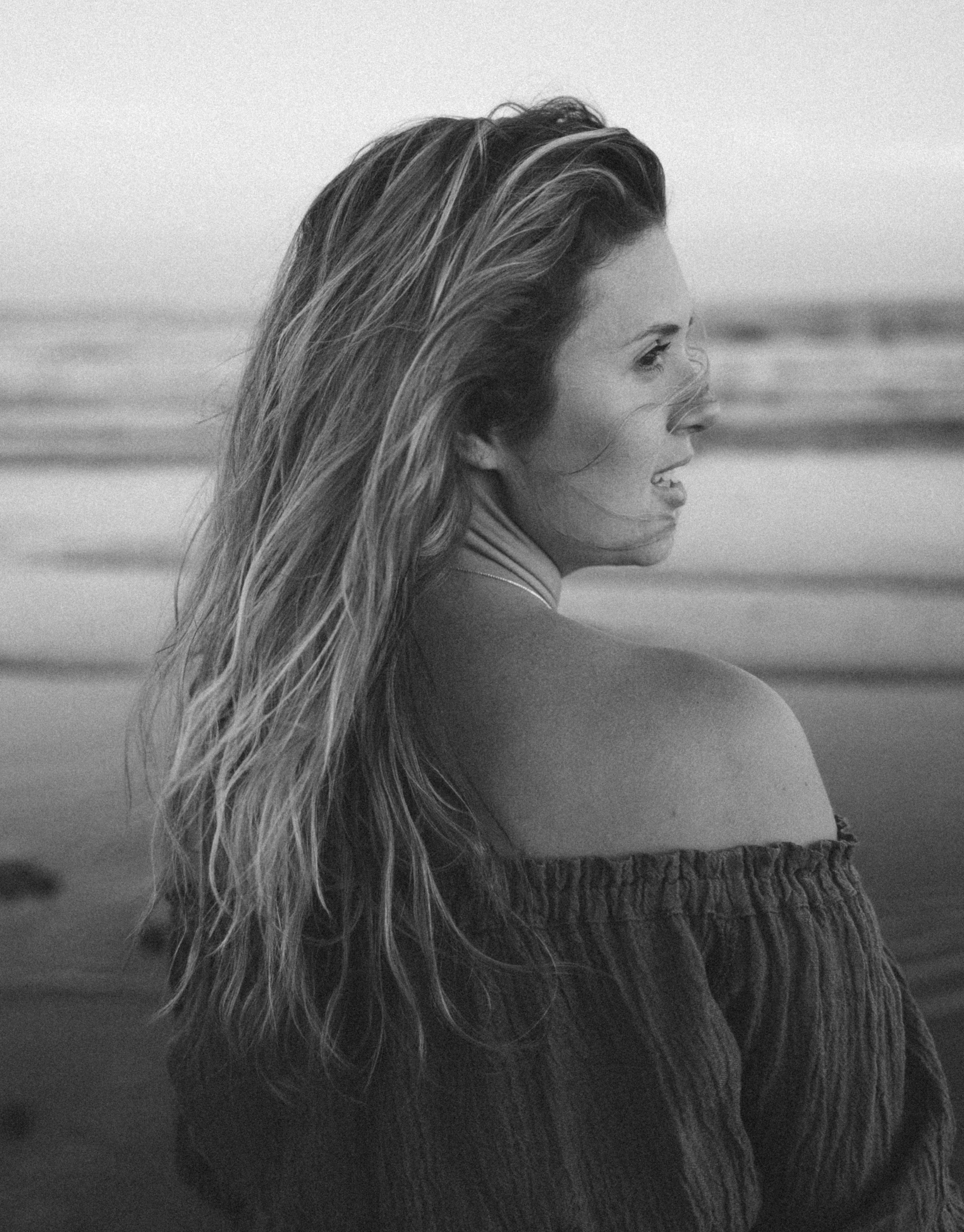Black and white photo of a woman with long, wavy hair looking to the side, standing on the beach near the ocean.