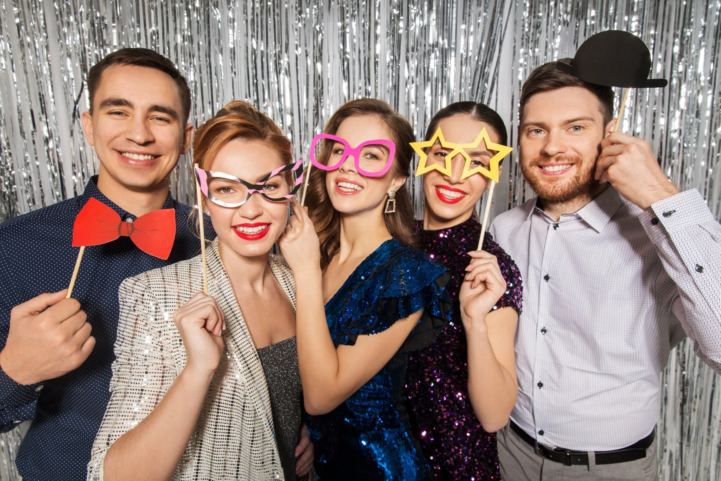 A group of five young adults dressed in party attire, holding playful photo booth props including glasses, bow tie, and hats, smiling in front of a silver tinsel backdrop.