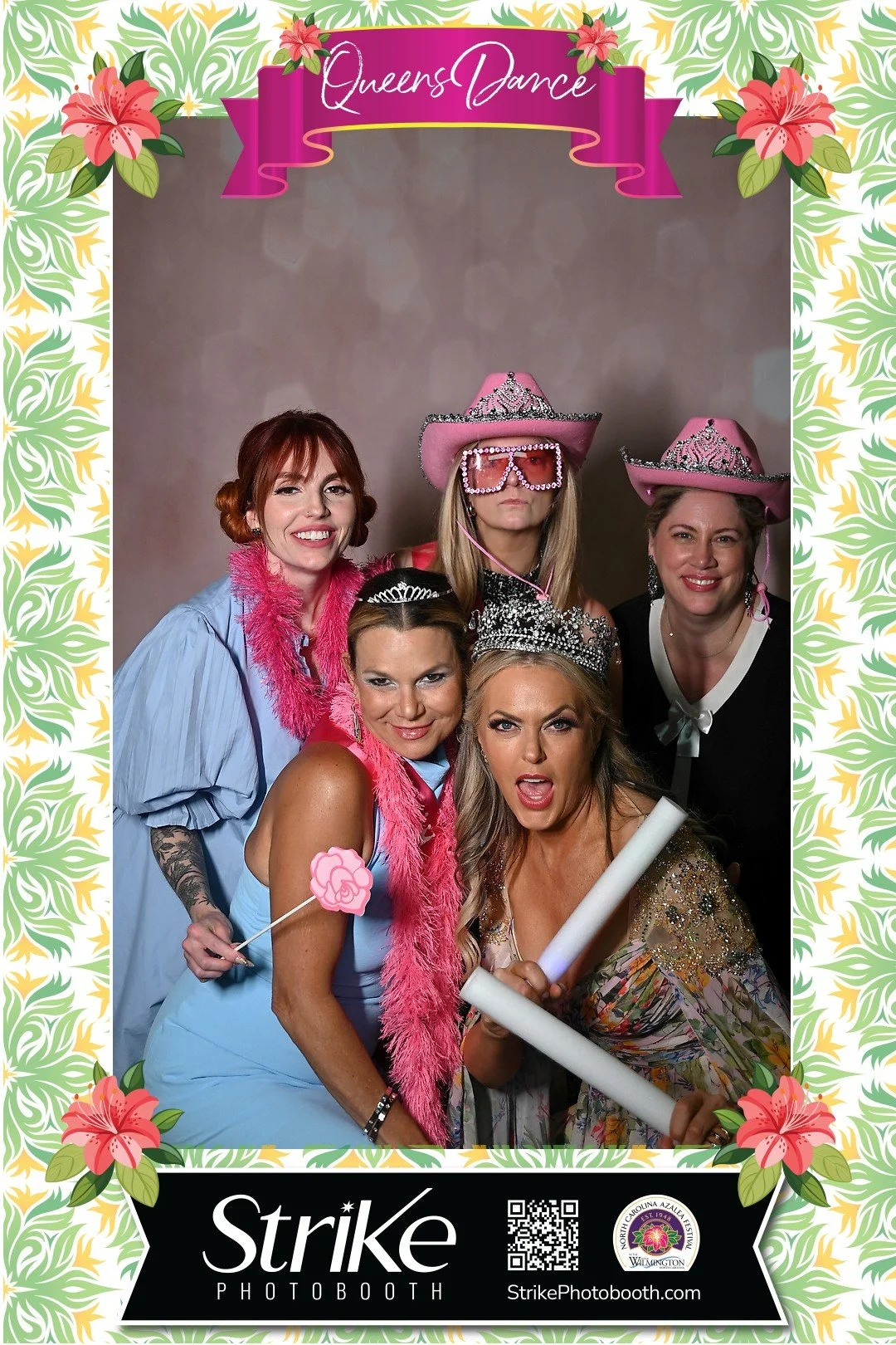 Group of five women dressed up for a party in a photo booth, wearing tiaras, pink feather boas, and fun accessories like pink sunglasses, cowboy hats, and holding props such as a pink flower and rolled papers. The photo is framed with a floral border and a banner that says 'Queen's Dance,' with a logo and QR code at the bottom.
