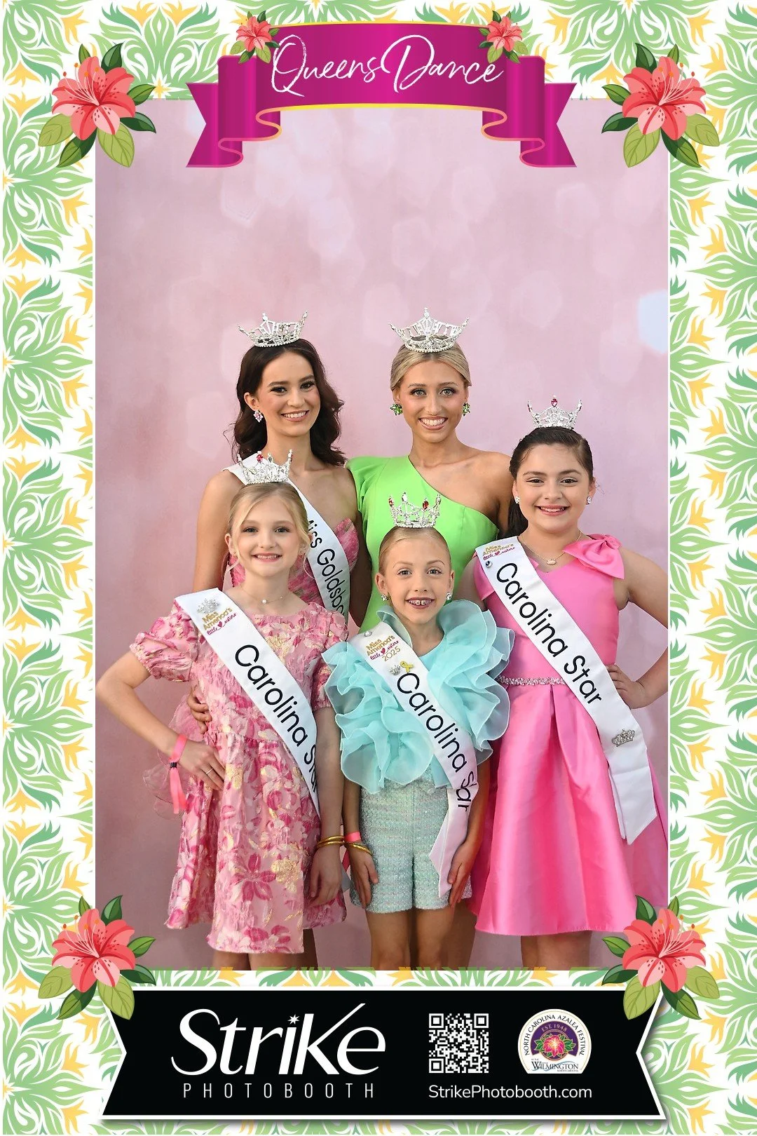Group of five young girls and two women dressed in colorful dresses and tiaras, standing in front of pink backdrop. The girls and women are wearing sashes and crowns, indicating a pageant or beauty contest event.