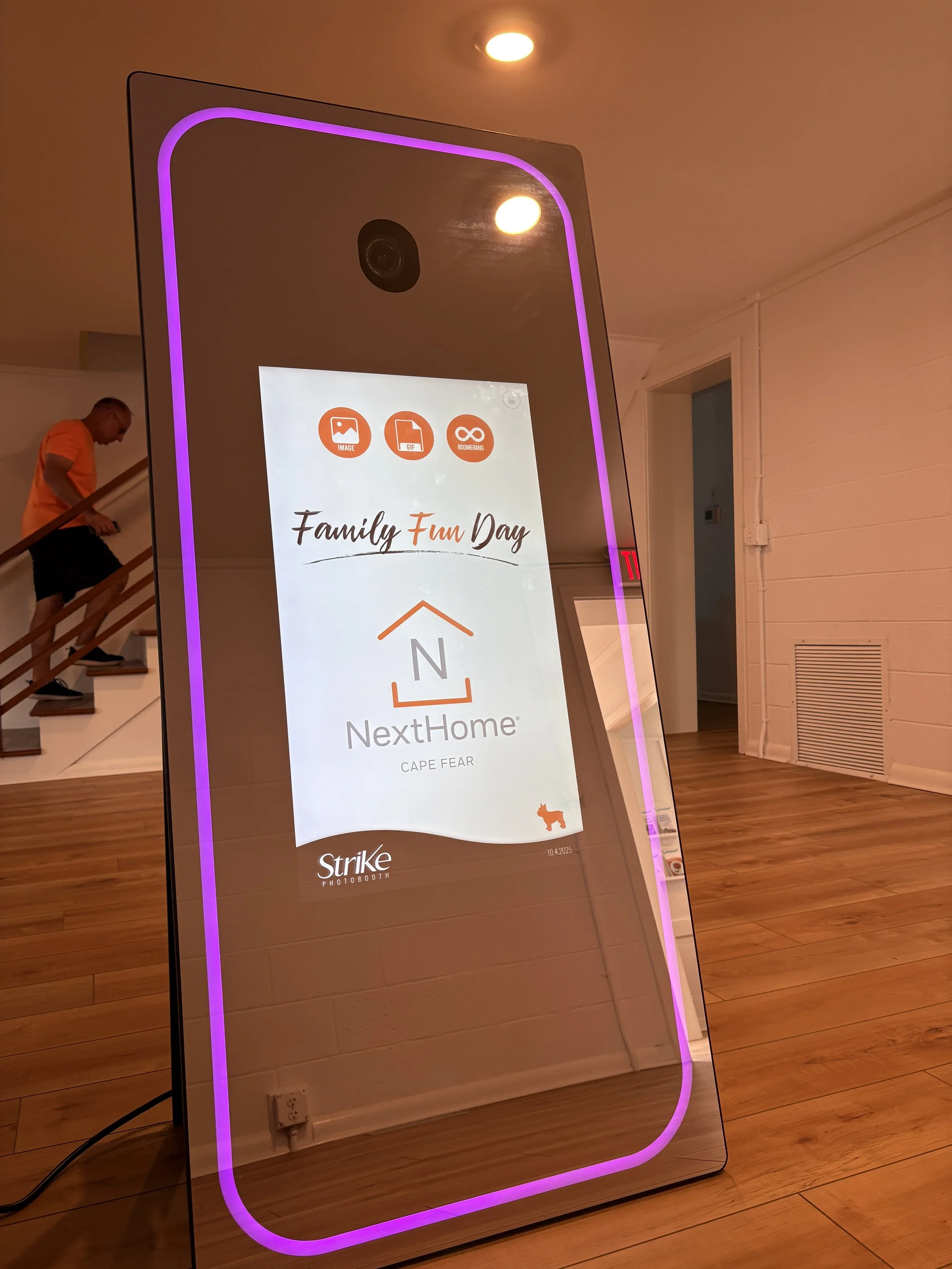A large, vertical, mirror with purple LED lighting around the edges. The reflection shows a wooden staircase with a man in an orange shirt. The mirror displays a digital advertisement for NextHome, a real estate company, featuring the message 'Family Fun Day' and icons for image, GIF, and boomerang options. The setting appears to be inside a modern home or office with wooden flooring and white walls.