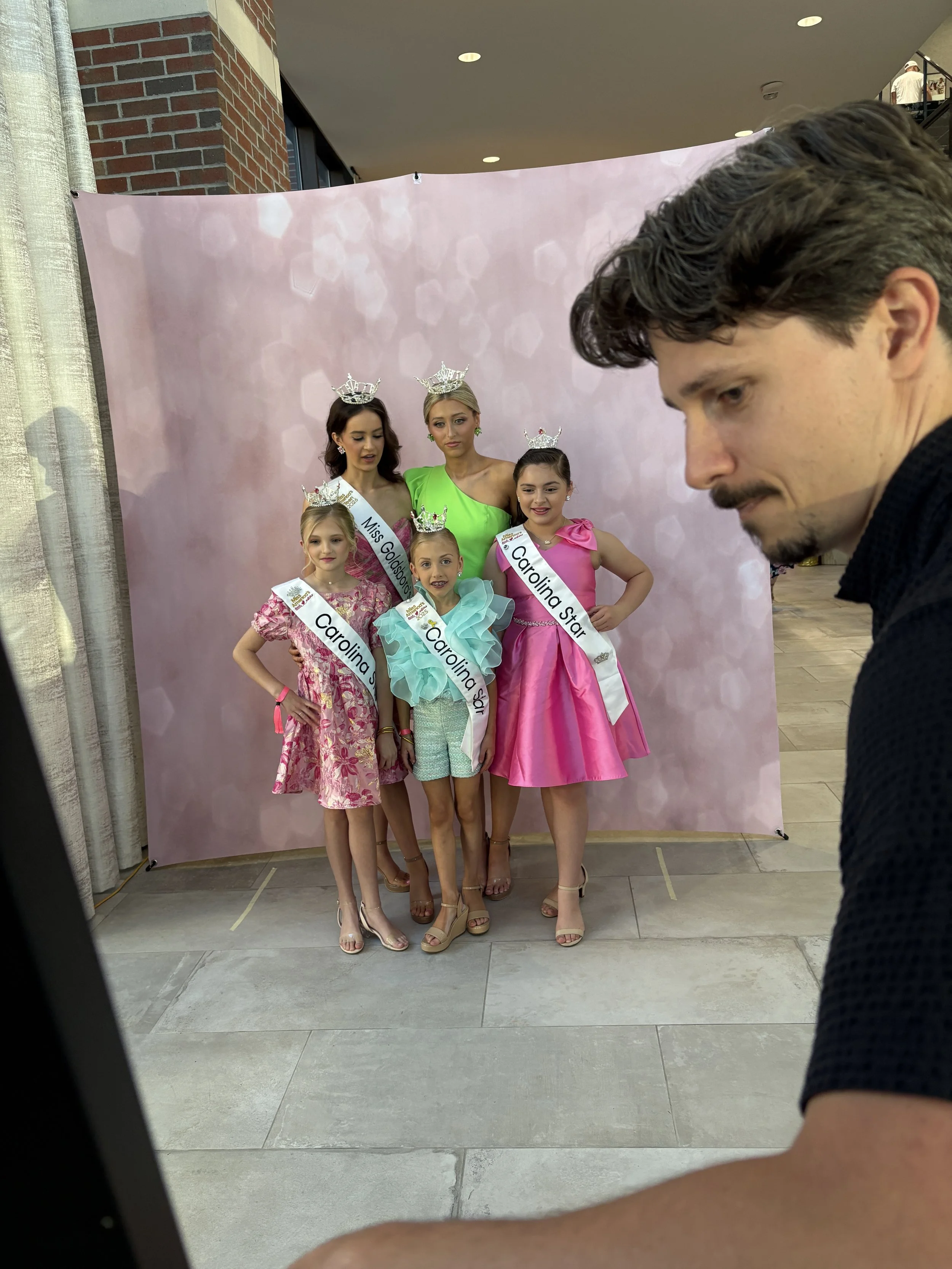 A group of five young girls and one woman, all wearing crowns and sashes that say 'Miss Goldsboro' and 'Carolina Star,' standing together for a photo in front of a pink backdrop. The girls are dressed in colorful dresses, and the woman is wearing a green dress. A man and another person are partially visible in the foreground and background, respectively.