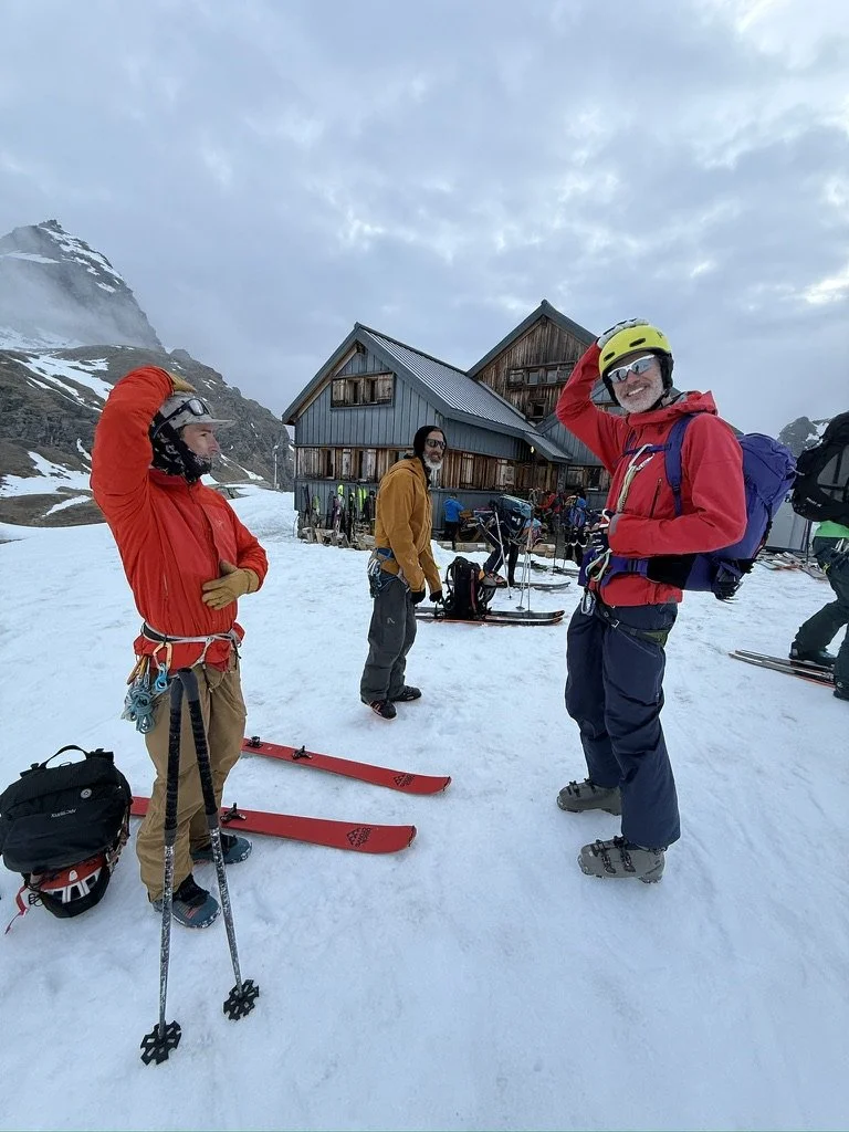 Group of skiers in winter gear standing on snow outside a mountain lodge, with skis and mountain scenery in the background.