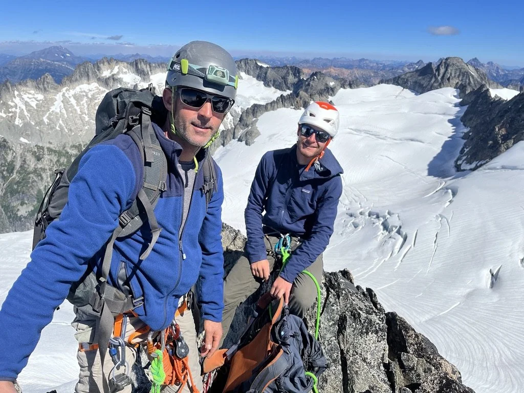 Two climbers in mountain gear standing on a snow-covered ridge with mountain peaks in the background