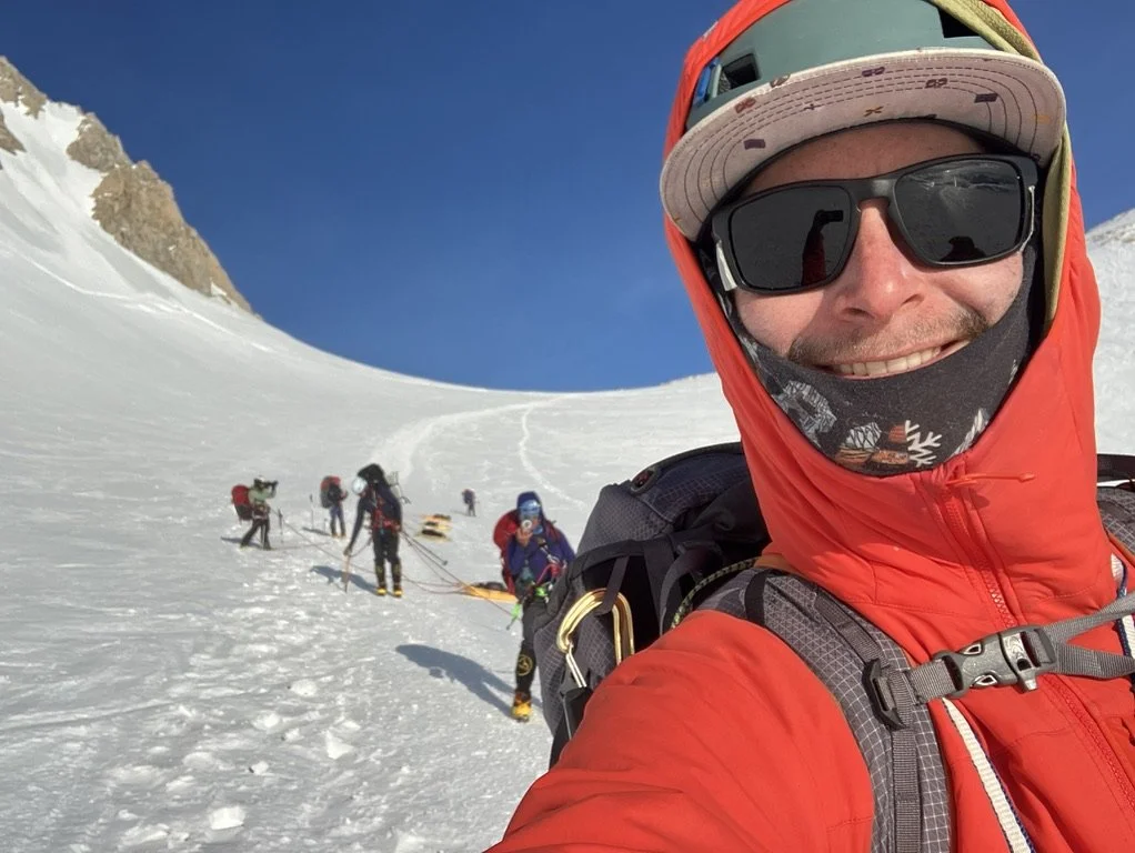A man taking a selfie on a snowy mountain with a group of climbers behind him, all wearing winter gear and sunglasses, under a clear blue sky.