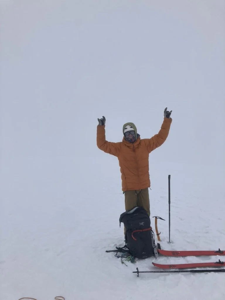 Person wearing orange jacket, goggles, and gloves standing in snow with arms raised, skiing equipment and backpack on the ground.