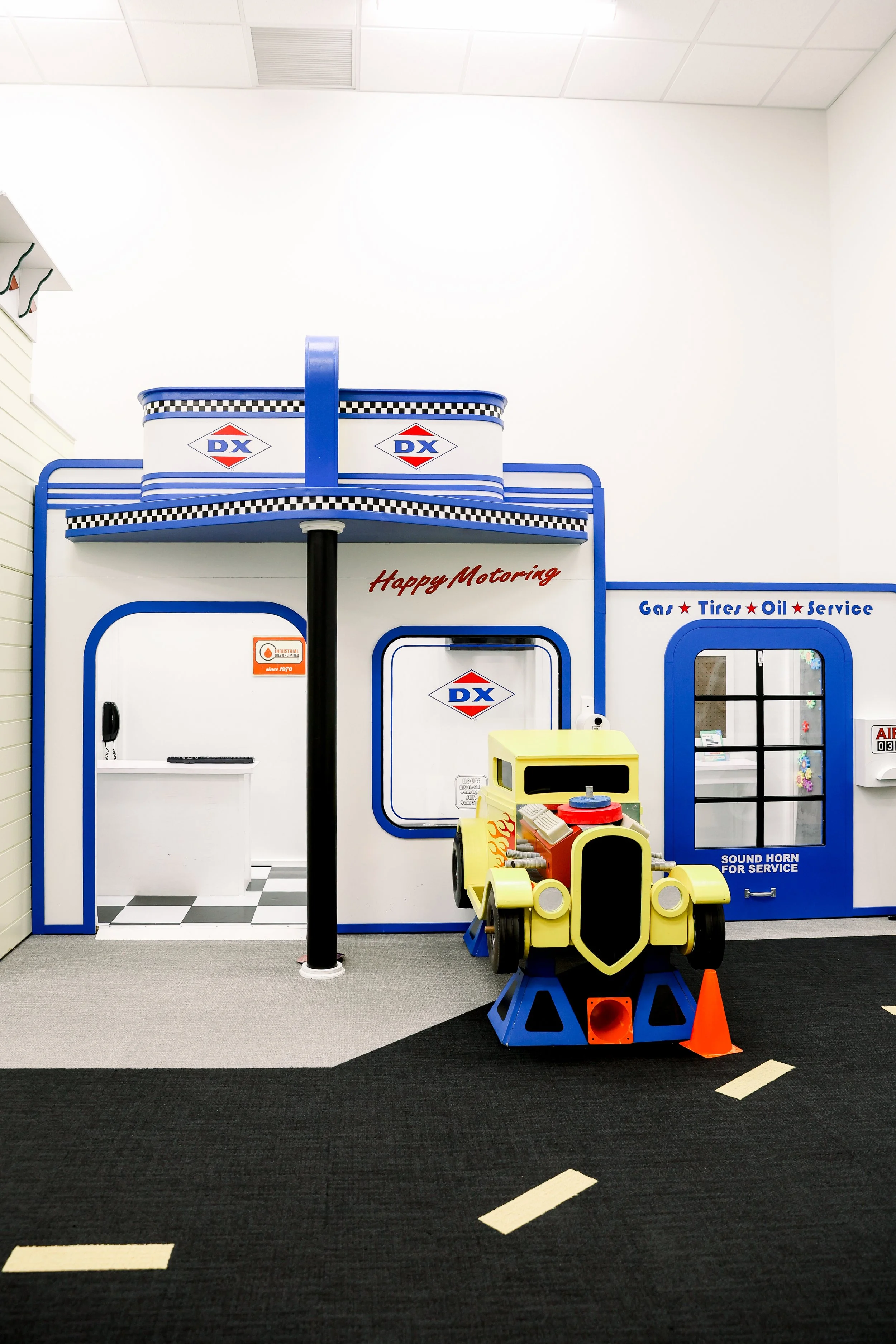 Kids pretend gas station with toy cars, gas pumps, and a yellow and black toy vehicle in an indoor play area.