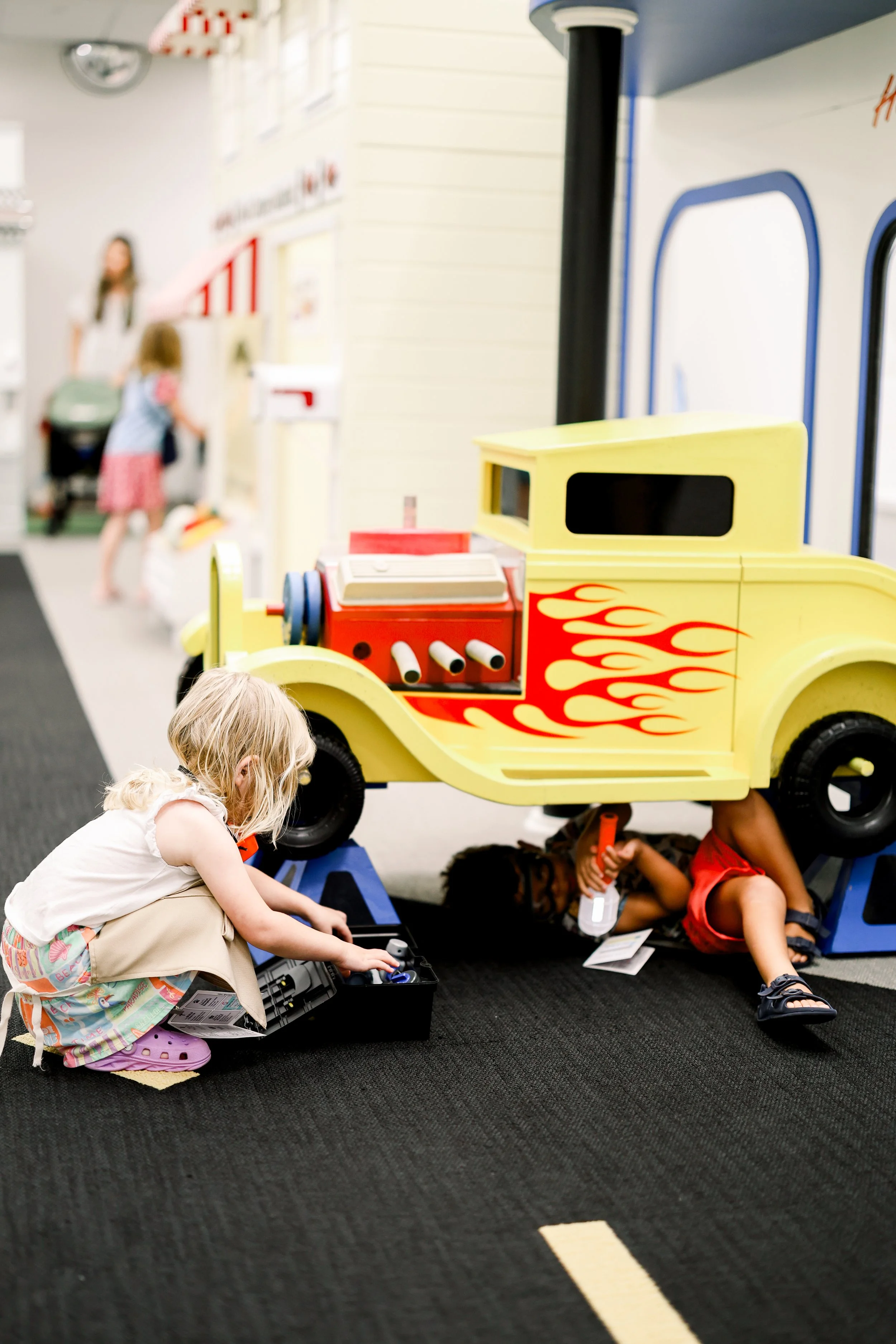 Children at a pretend ice cream shop with a pink counter, ice cream cones, and menu, including a girl with a red bow and a boy wearing glasses
