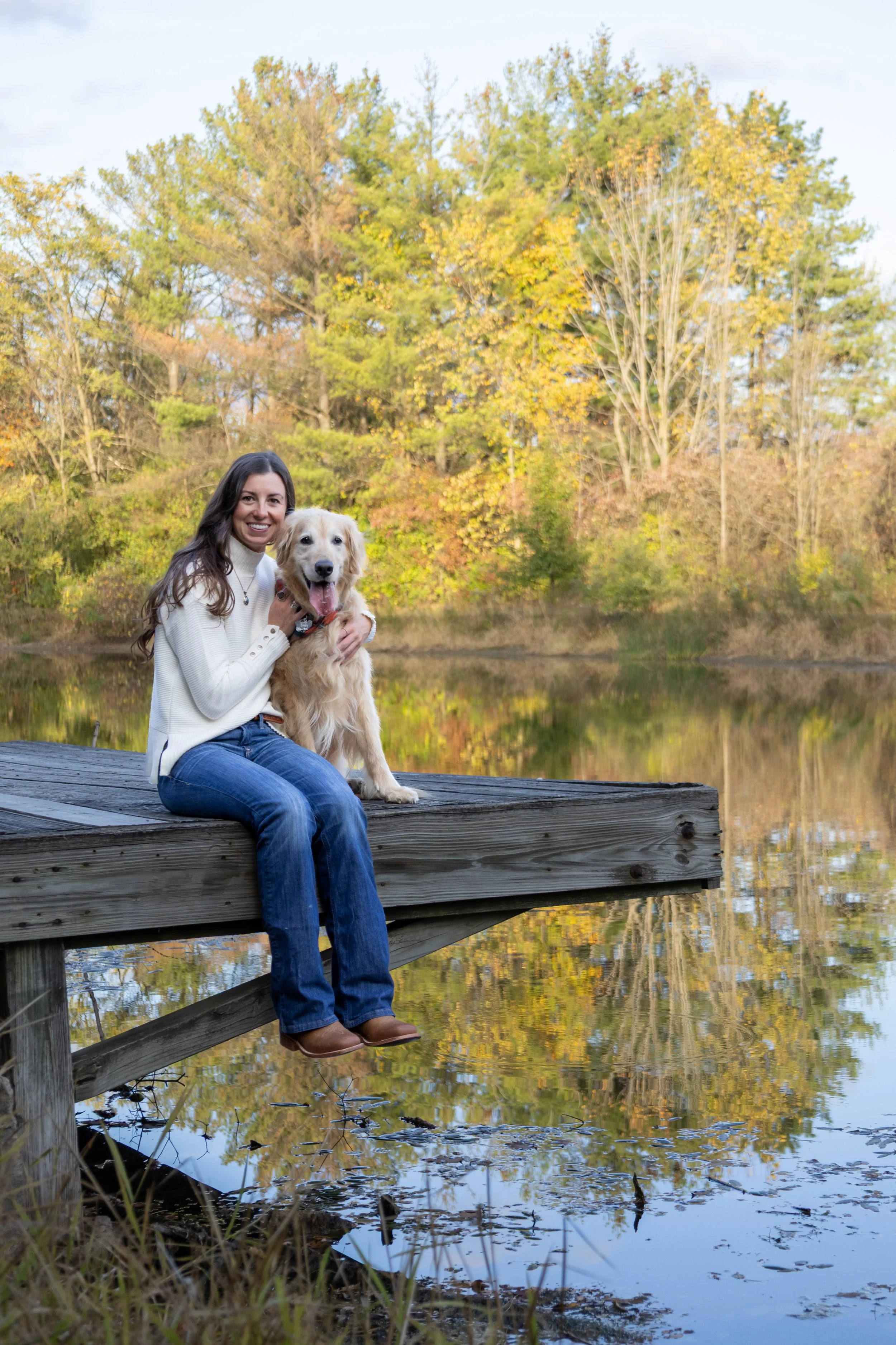 dog and mom sitting together at a dock in a homestead park in fort wayne photographed with trees and pond behind them