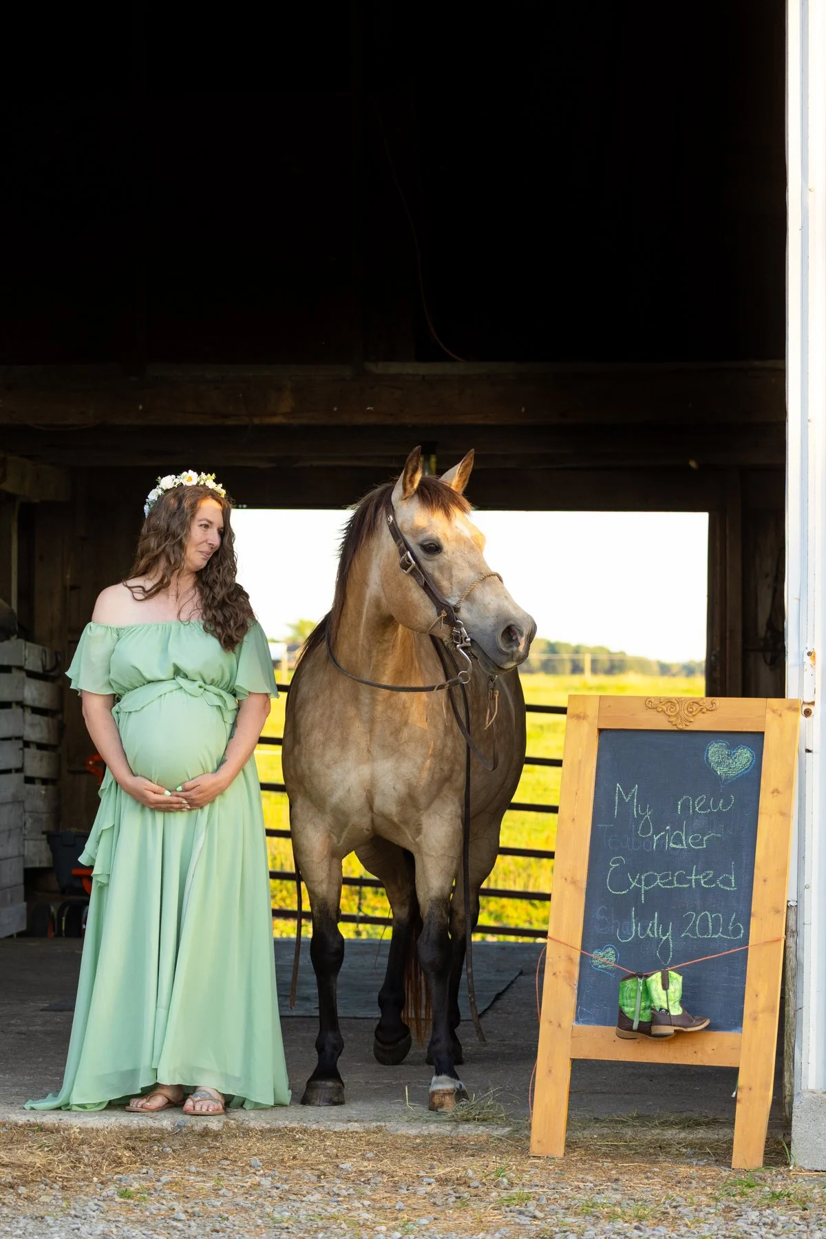 Maternity photo session with an expecting mom wearing a green flowy dress and flowers in her hair standing next to a horse in a barn with a sign that tells of the upcoming arrival of a newborn baby
