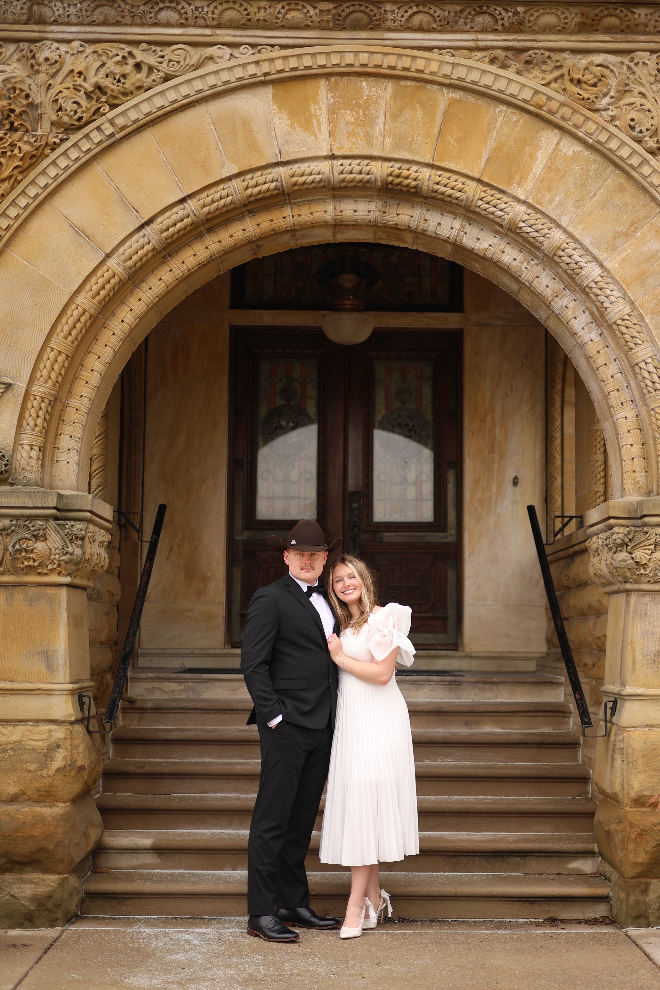 man and woman standing together and smiling at photographer in front of a beautiful historic building at st francis in fort wayne