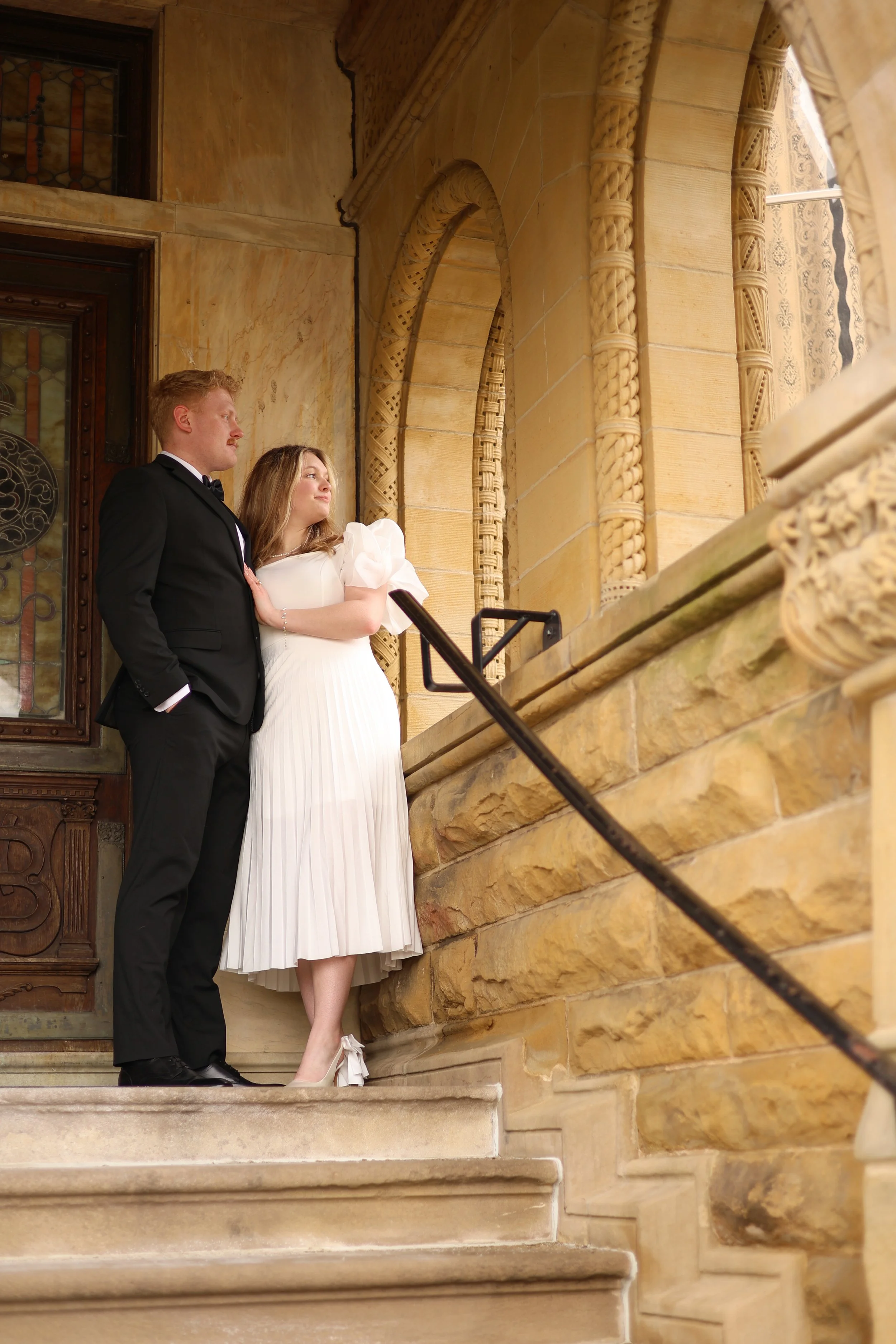 man and woman standing together on the steps of a historic building in fort wayne photographed from below and looking off the in distance and thinking of the wedding