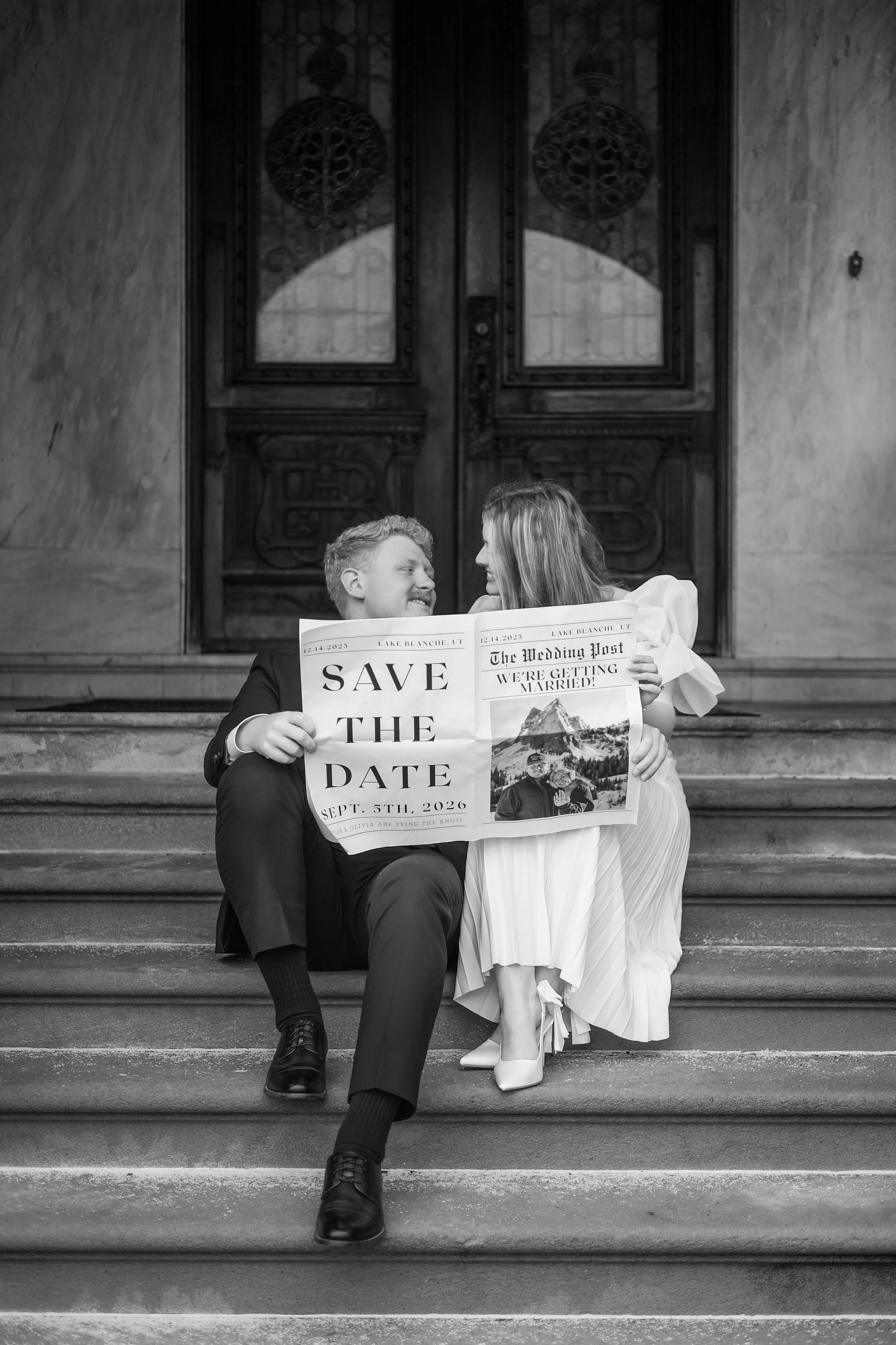 man and woman sitting on steps of a building at st francis at fort wayne reading a newspaper that is announcing the save the dates for the upcoming wedding photographed in black and white