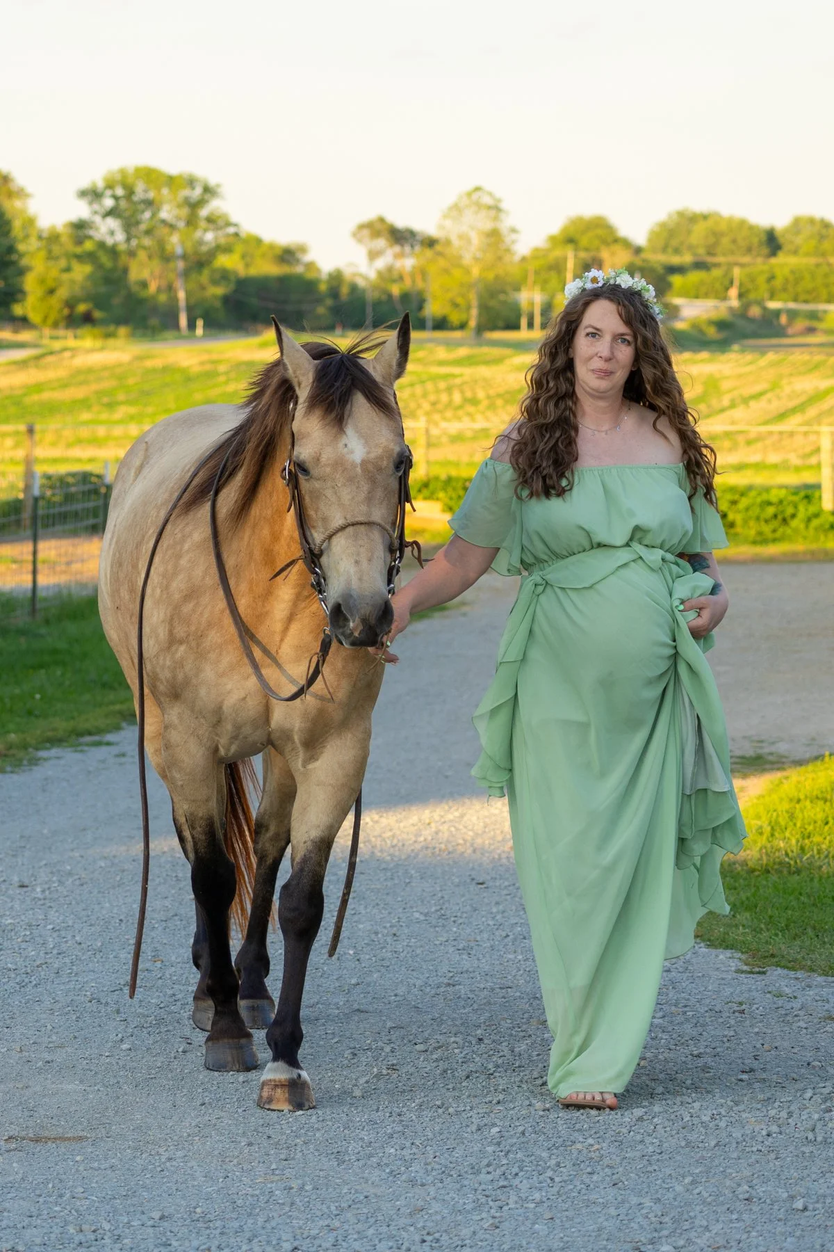 Maternity photography session at a farm in North Manchester, Indiana, with an expectant mom in a long green flowy dress with flowers in her hair walking next to a horse