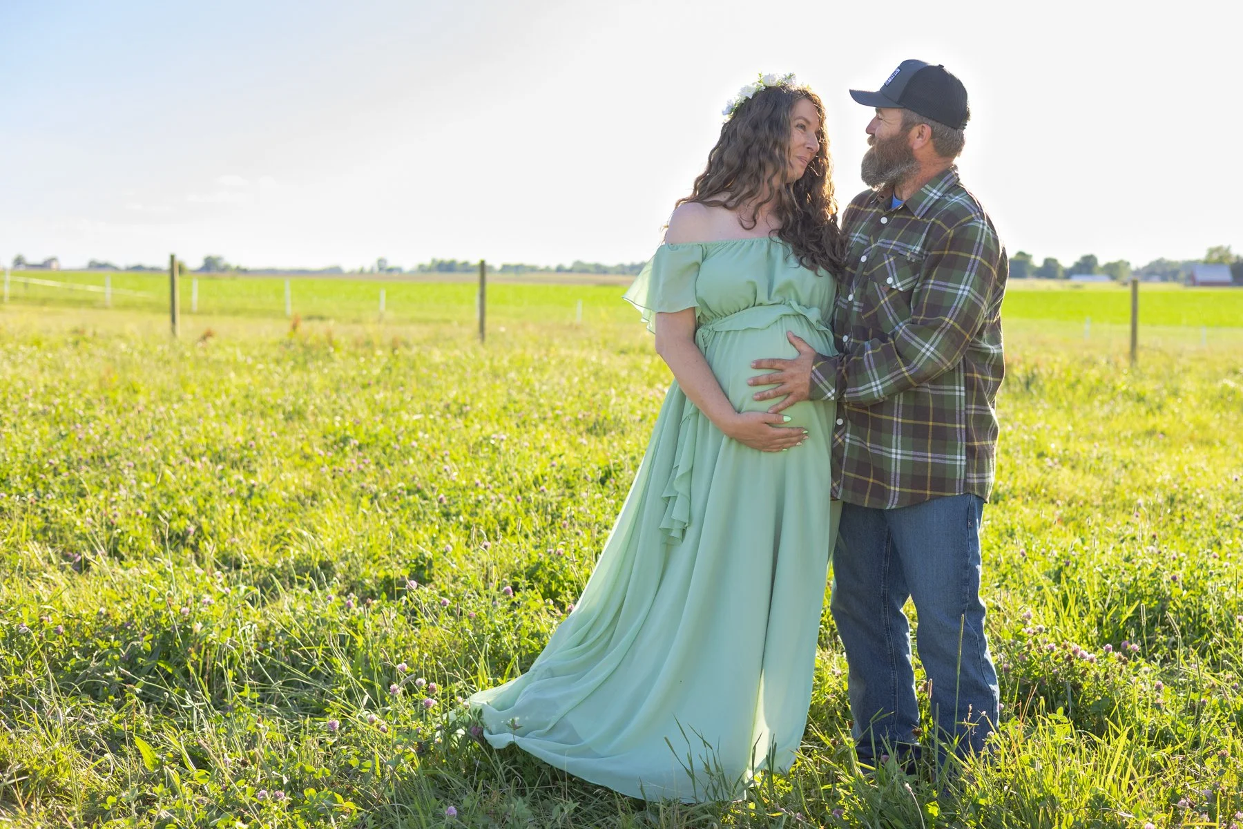 Dad and Mom holding onto Mom's belly as they are expecting a newborn soon, maternity photo with mom in a green dress taken in farmfields in Indiana