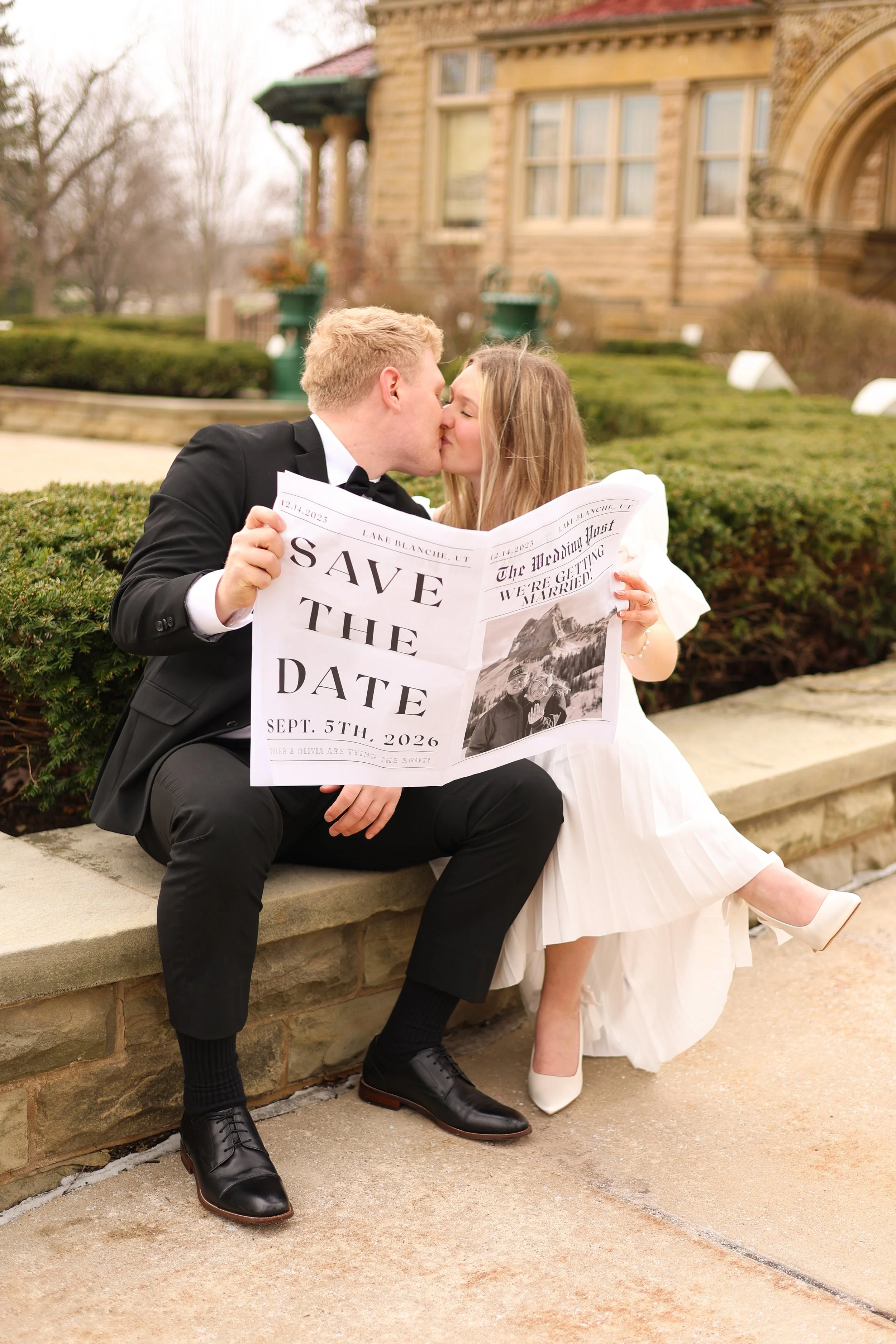 man and woman kissing while holding a newspaper that announces the wedding in the save the dates photograph taken at st francis in fort wayne