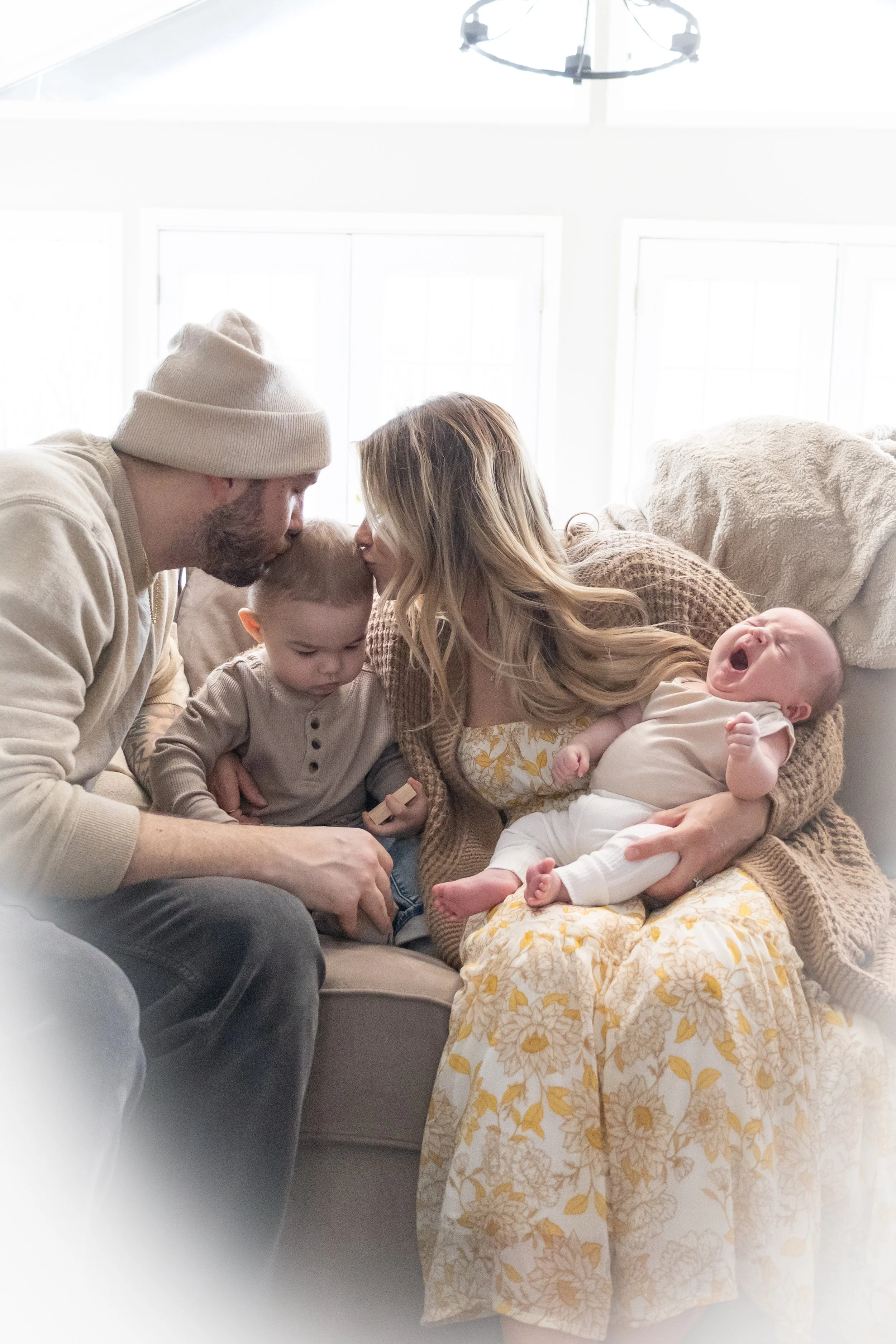 Family with new baby photograph in home in Huntington Indiana the mom and dad are kissing the toddler and the baby is resting in mom's arms
