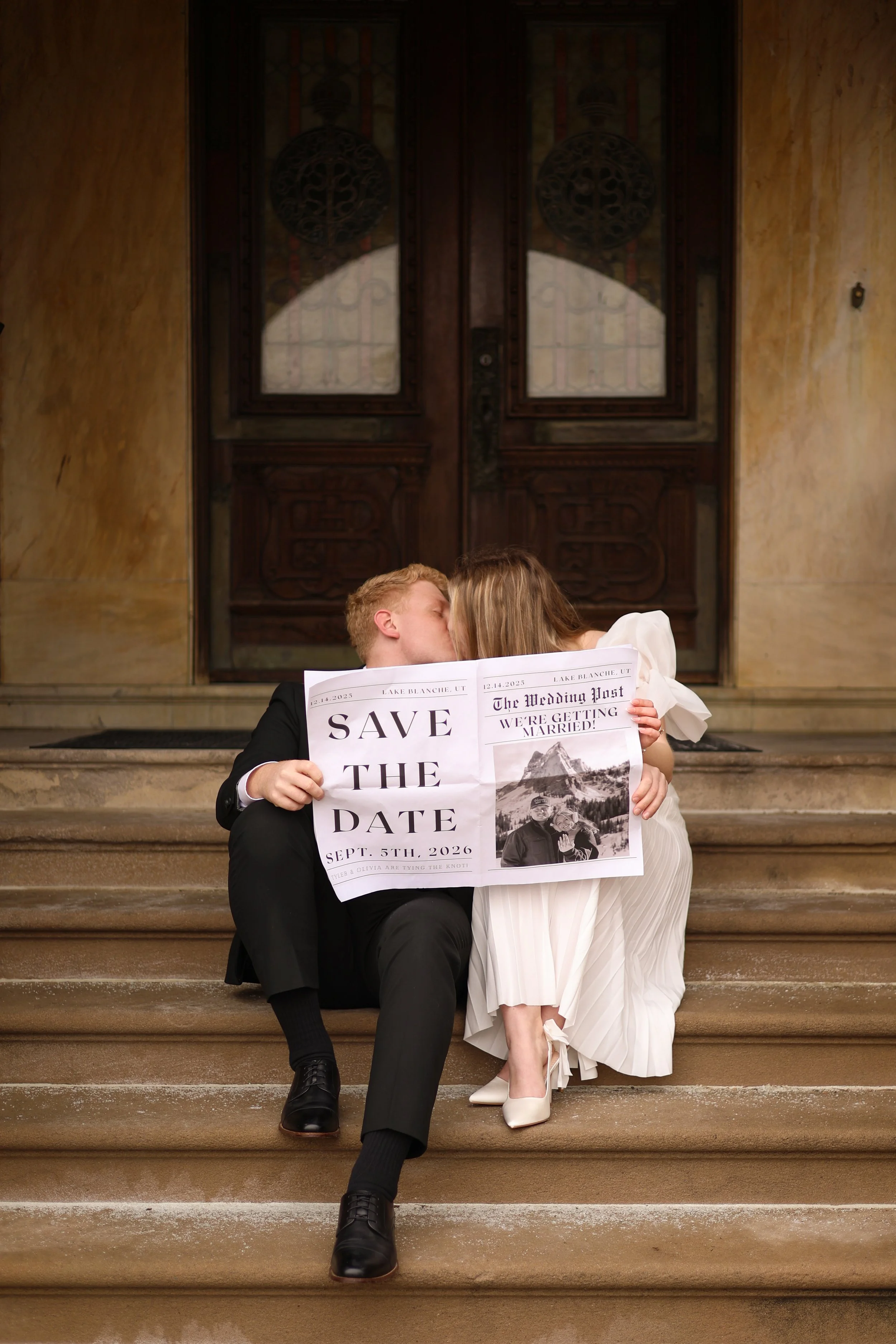 man and woman kissing on the steps of a historic building at st francis in fort wayne they are reading a newspaper to announce their save the dates for the upcoming wedding photographed to spread the word