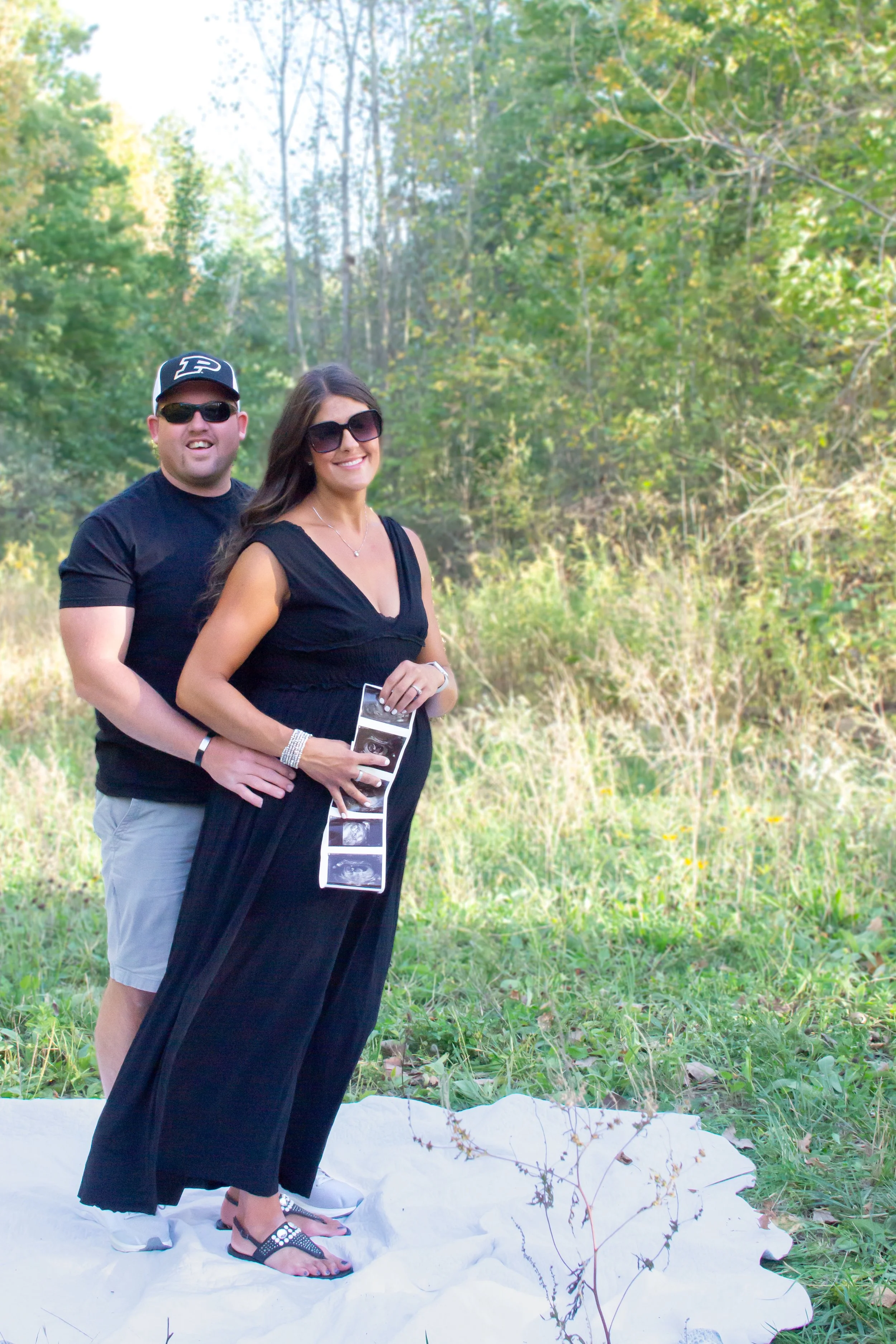 New Dad and Mom standing together and smiling for joy with the ultrasound iamges of their new baby at park with trees in Huntington Indiana