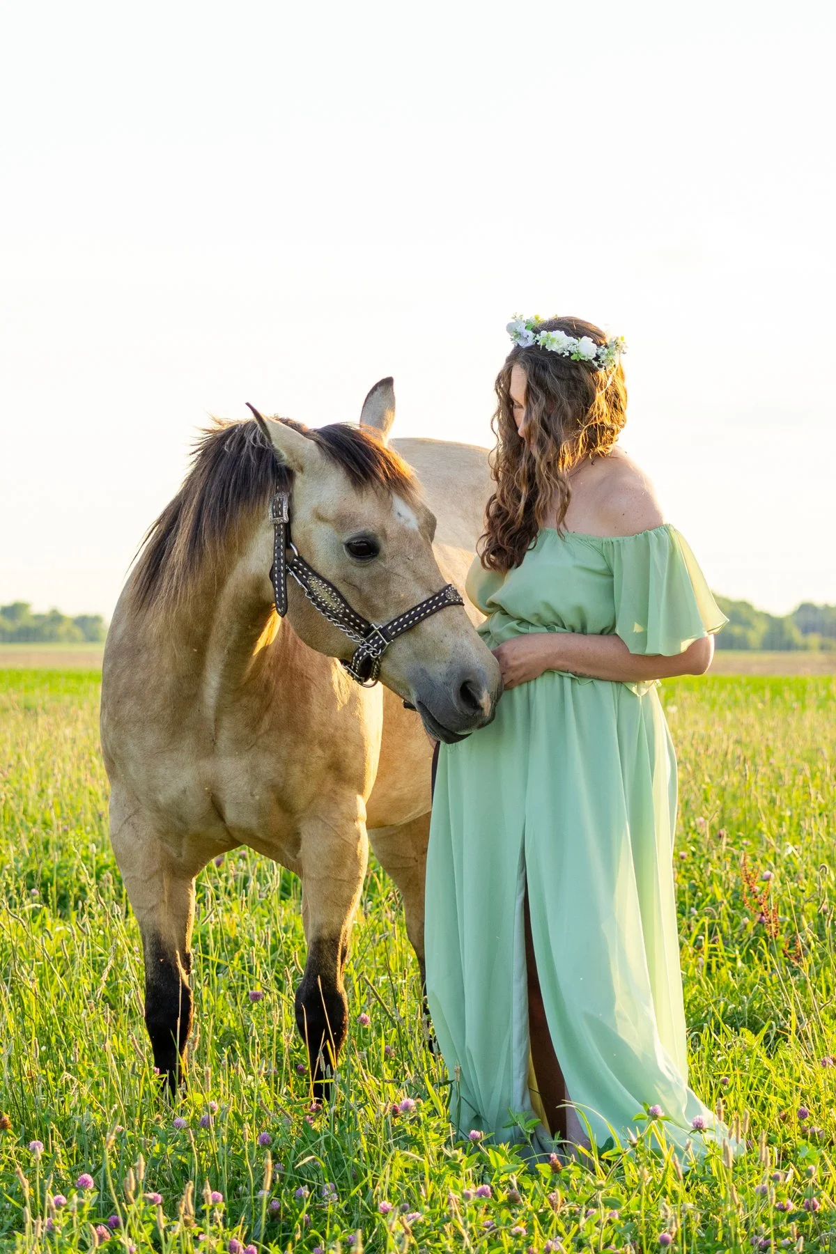 Maternity photo taken of expectant mom in a green flowly dress with flowers in her hair holding onto her horse who is nudging her belly photographed in an Indiana grassy farm field at sunset
