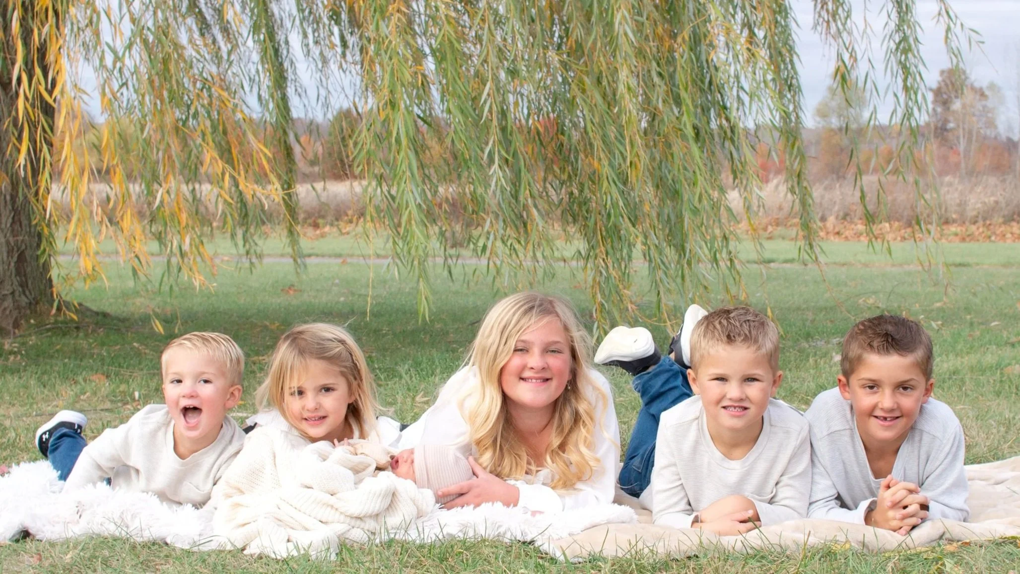 Family under a willow tree laying on a blanket under a willow tree with a newborn and lots of brothers and sisters photographer captured them smiling at the camera