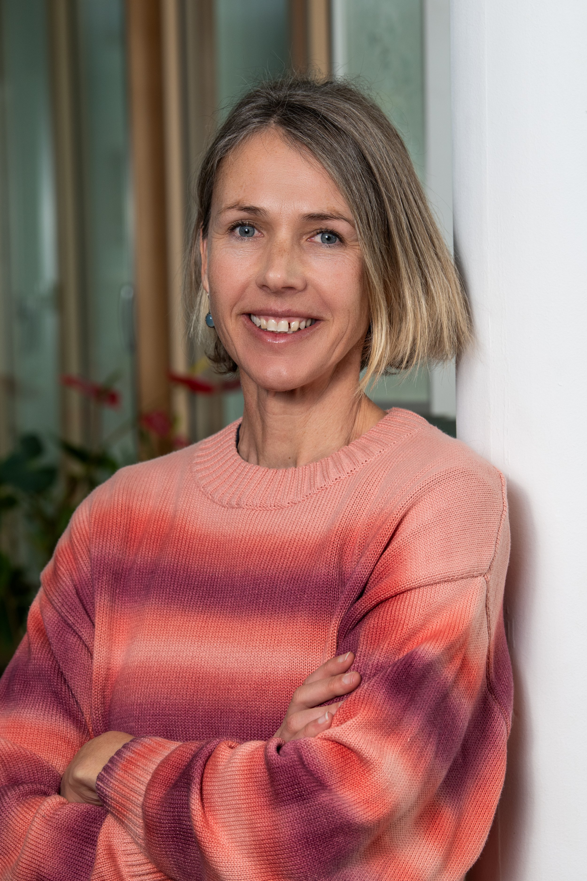 Eine lachende Frau mit kurzen, blonden Haaren trägt einen pinken Pullover mit Streifen und steht vor einer weißen Wand. Im Hintergrund sind Fensterrahmen sichtbar.