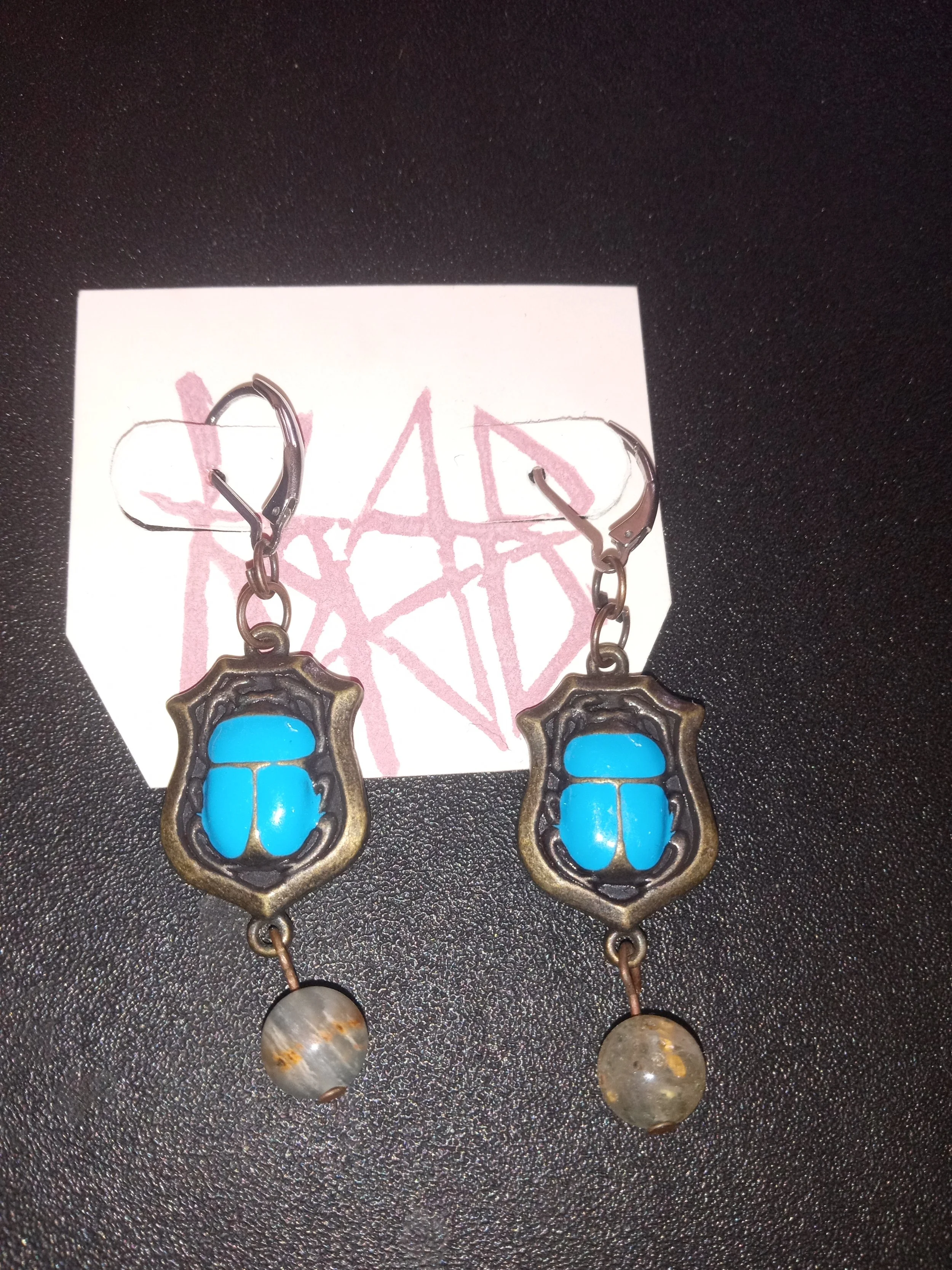 Pair of earrings with turquoise beetle design, attached to a white card with pink text, on a black surface. Artisan, Handmade, Grass valley, nevada county, Kale Burnett Earrings, art.
