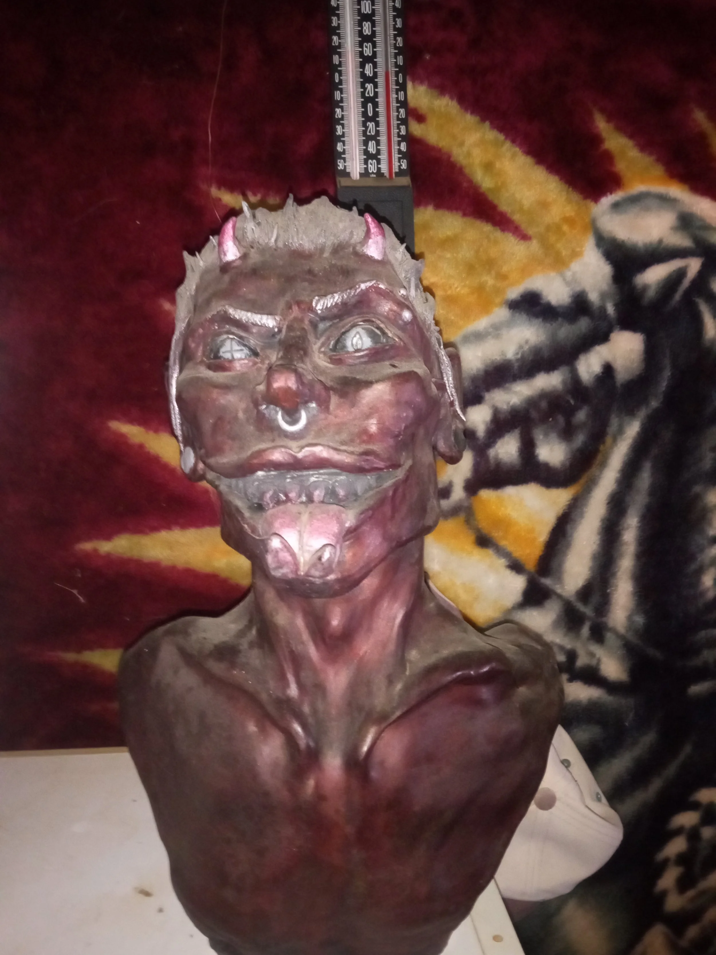 A sculpture of a humanoid face with animalistic features, including horns, fangs, and a nose ring, placed against a colorful background, with a measuring ruler above it. Artisan, Handmade, Grass valley, nevada county, Kale Burnett, Ceramic Sculpture