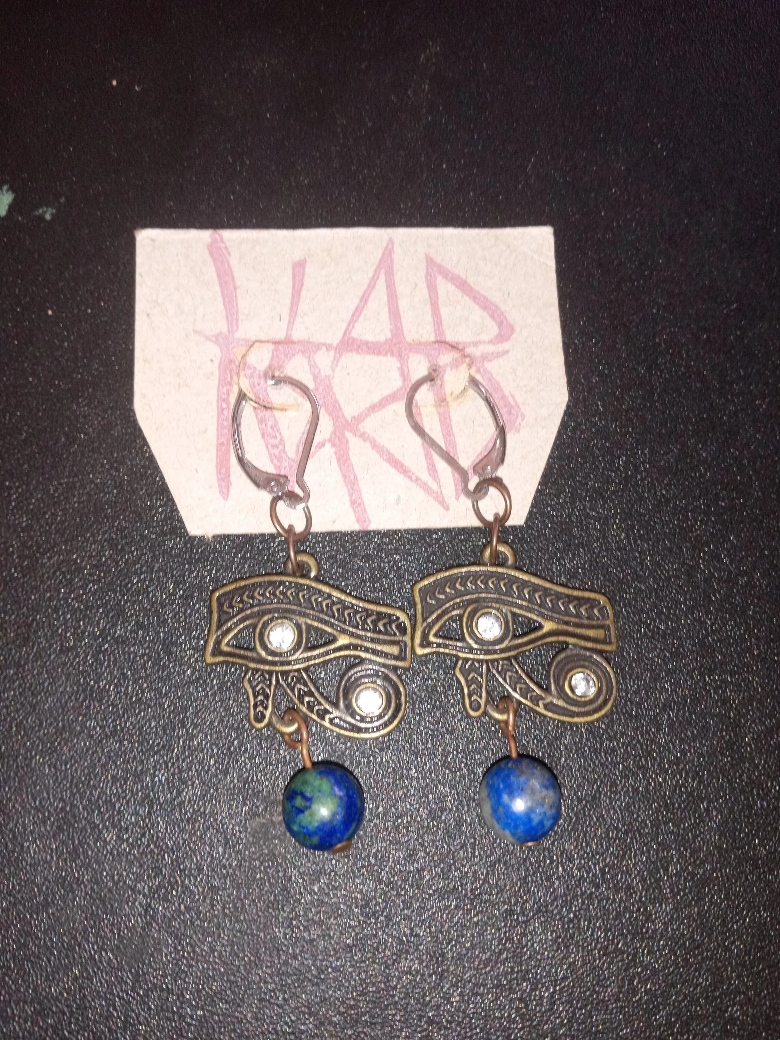 A pair of earrings with butterfly-shaped pendants and blue spherical beads hanging from a card with pink scribble writing. Artisan, Handmade, Grass valley, nevada county, Kale Burnett Earrings, art.