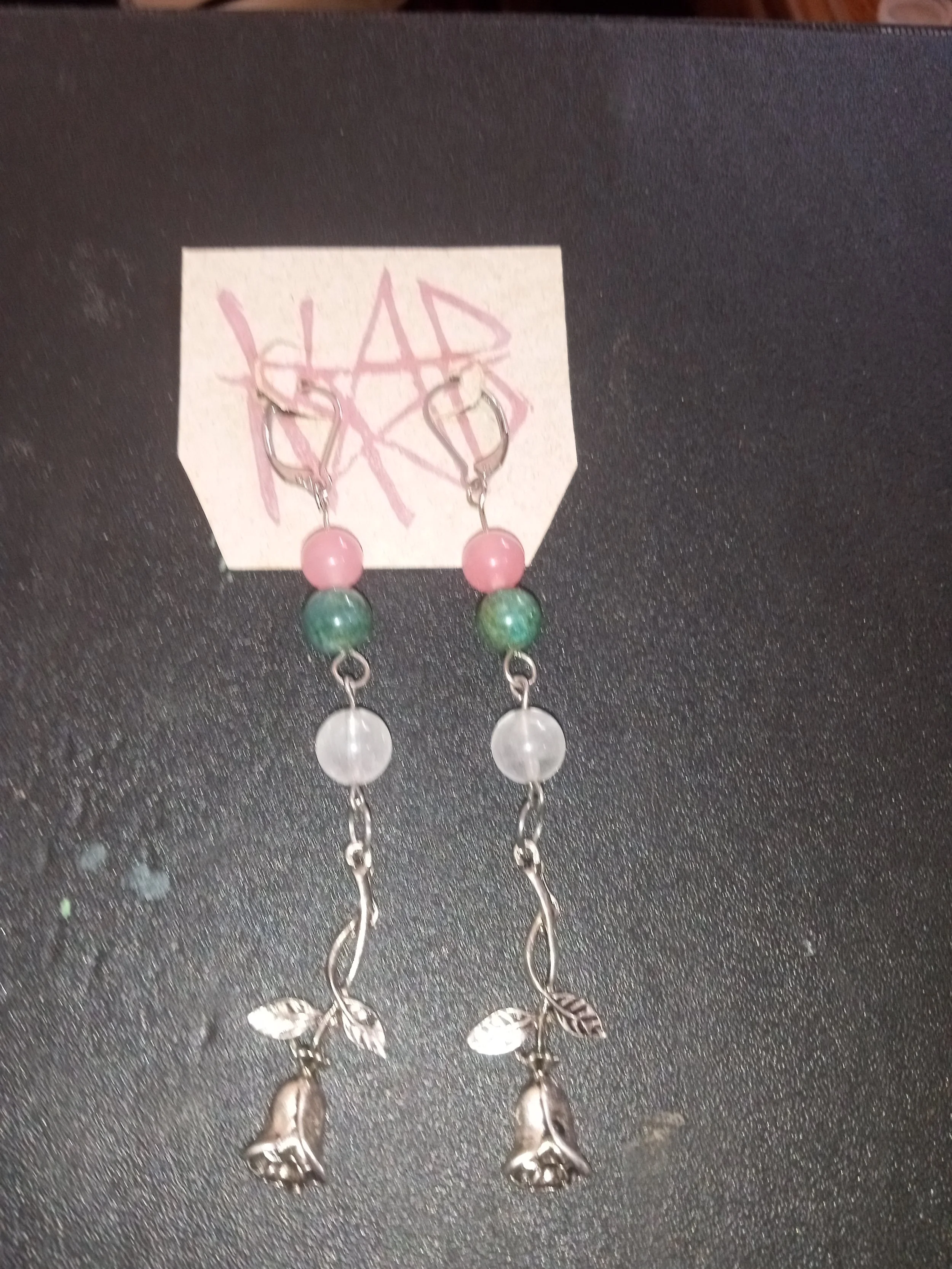 A pair of earrings with pink and green beads, shaped like branches with leaves, hanging on a black surface that has a handwritten note behind them. Artisan, Handmade, Grass valley, nevada county, Kale Burnett Earrings, art.