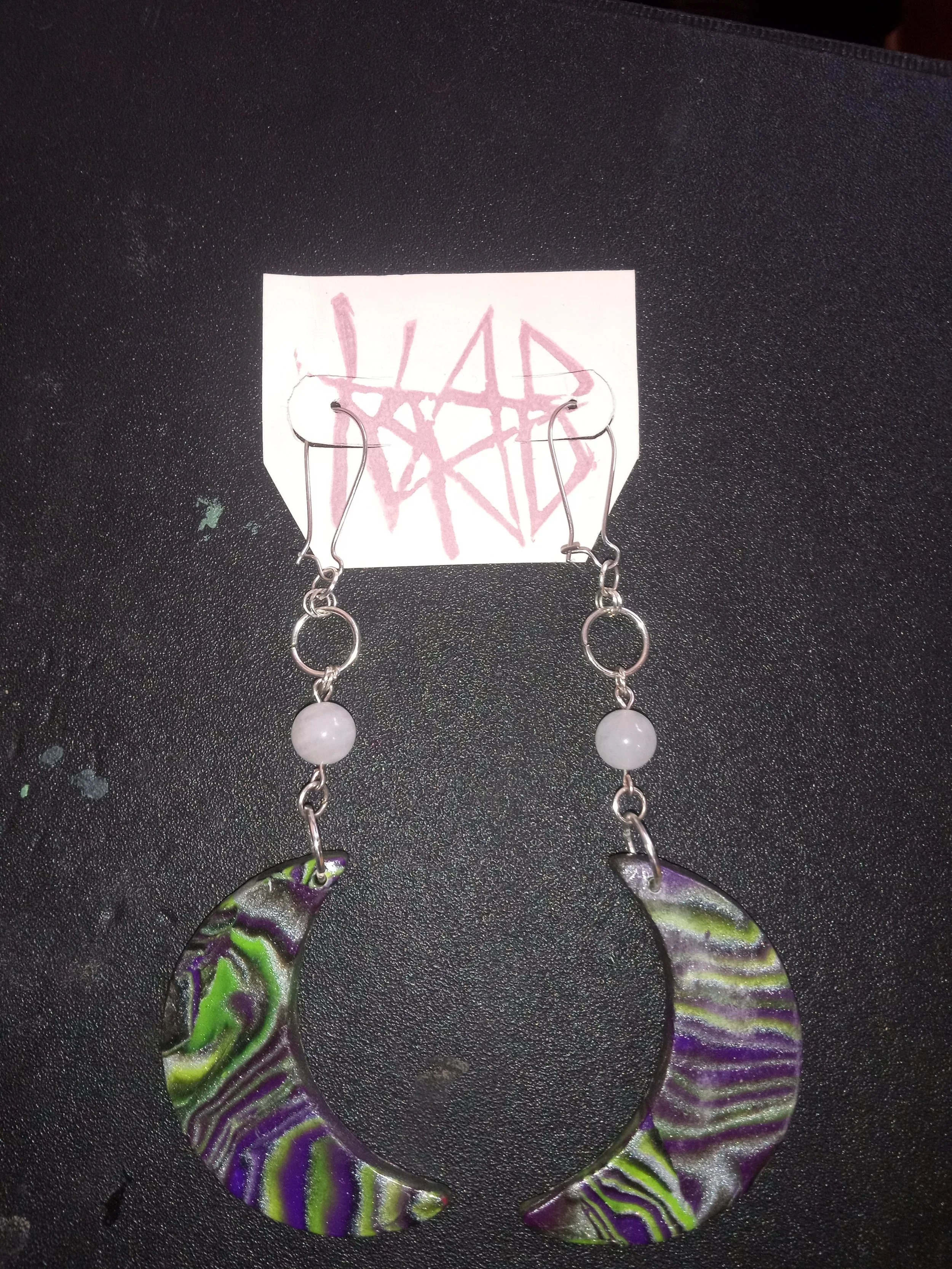 A pair of colorful, spiral-shaped earrings with purple, green, and yellow patterns hanging from a wire display on a black textured surface. Artisan, Handmade, Grass valley, nevada county, Kale Burnett Earings, art.