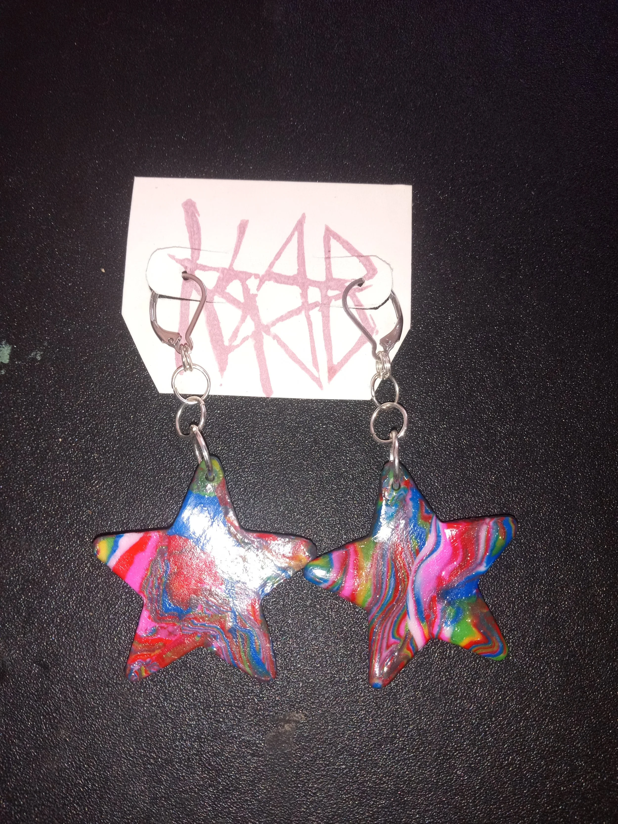 Colorful star-shaped earrings with a swirled rainbow pattern, hanging on a paper with red graffiti-style writing.Artisan, Handmade, Grass valley, nevada county, Kale Burnett Earrings, art.