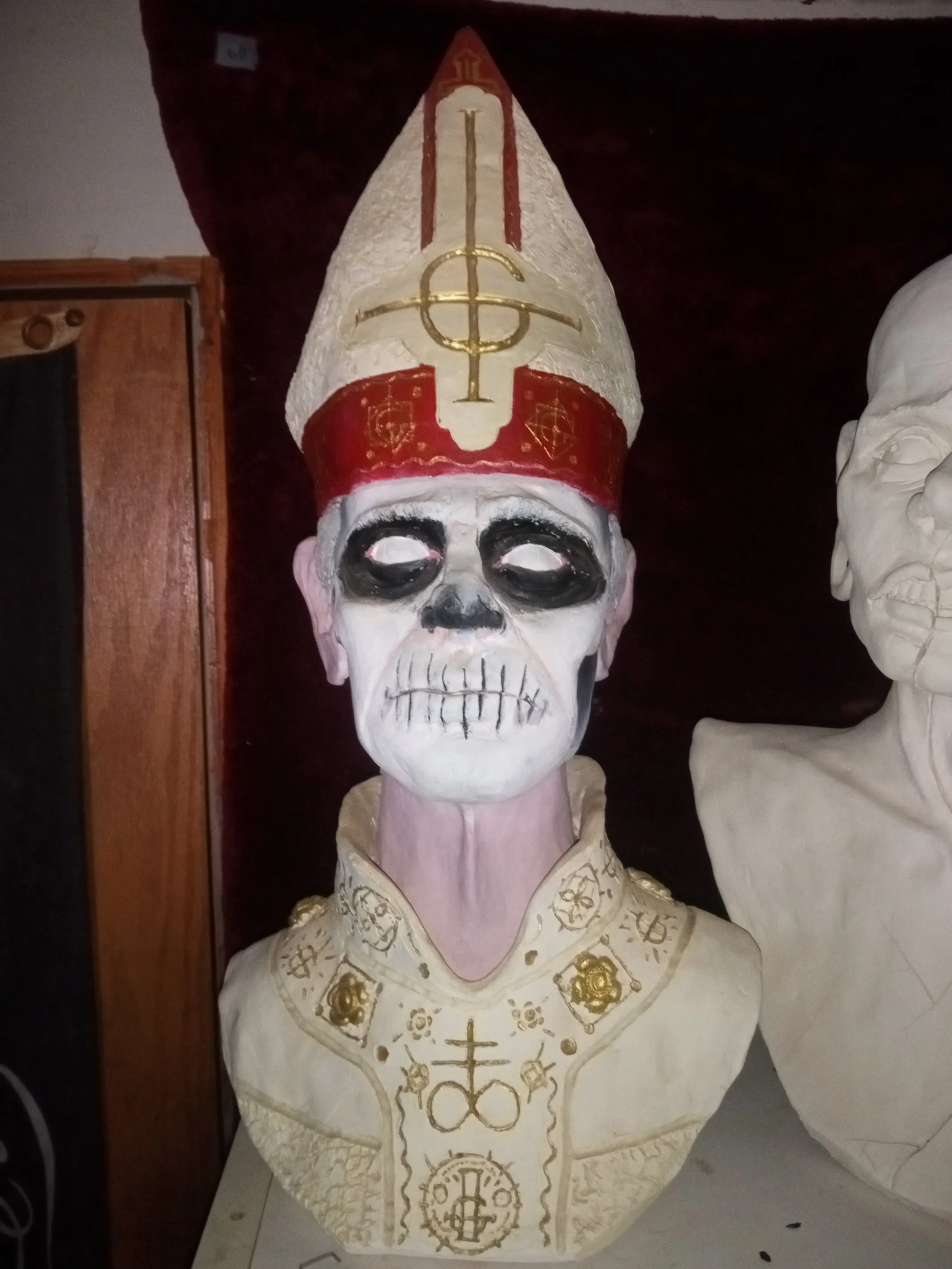 A skull sculpture painted white with gold and red symbols and designs, wearing a tall, pointed mitre with religious symbols. Artisan, Handmade, Grass valley, nevada county, Kale Burnett, Ceramic Sculpture of "Ghost's Papa Nihil" Character.