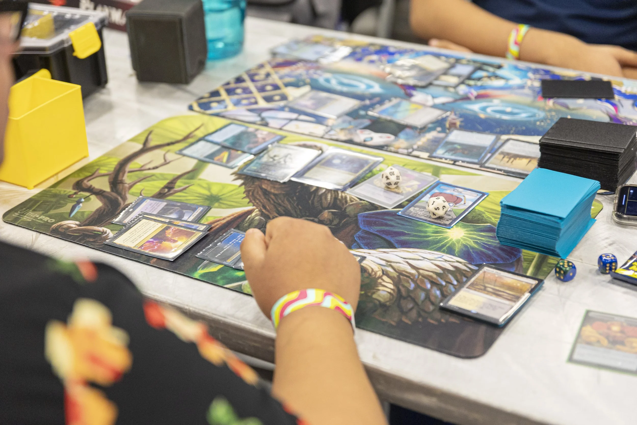 People playing Magic: The Gathering card game at a table with colorful playmat, various cards, dice, and card stacks.