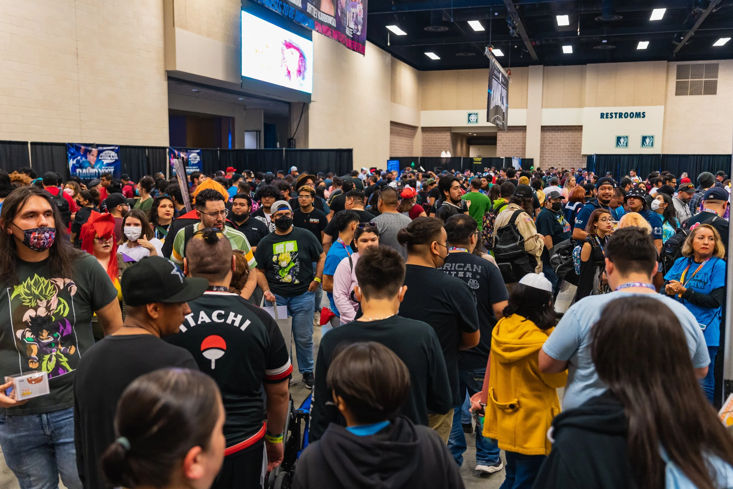 Crowd of people at a convention with some wearing masks and costumes, gathered in a large indoor hall. There's a sign for restrooms in the background.