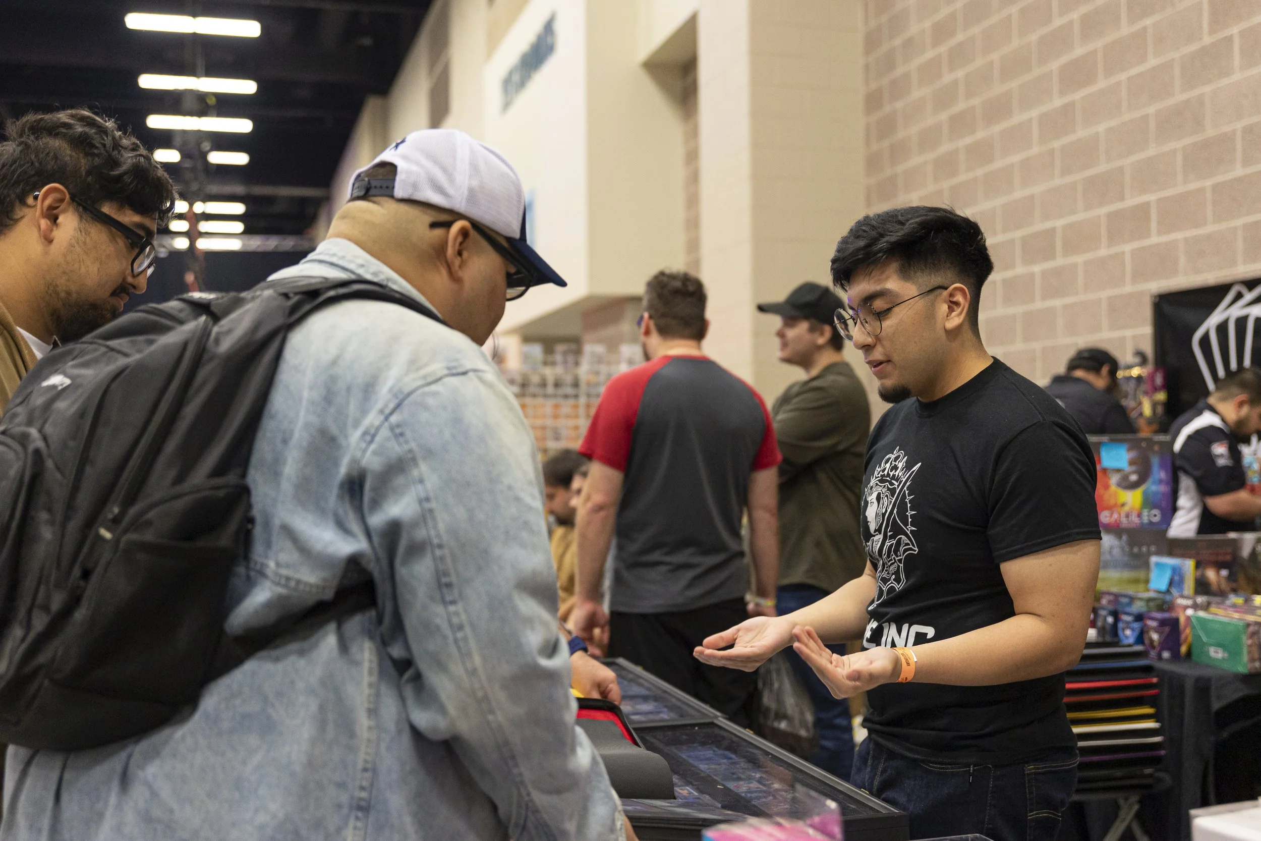 A man in black T-shirt demonstrating a collectible card game to a group of three men, one wearing a light denim jacket and cap, at a convention or gaming event.