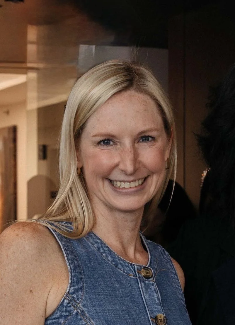 A woman with blonde hair wearing a sleeveless green top, smiling, against a brick wall background.