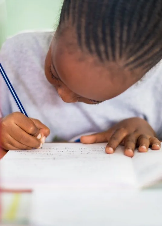 Clost up photo of a child with tight braids leaning down close to their paper writing with a blue pencil.