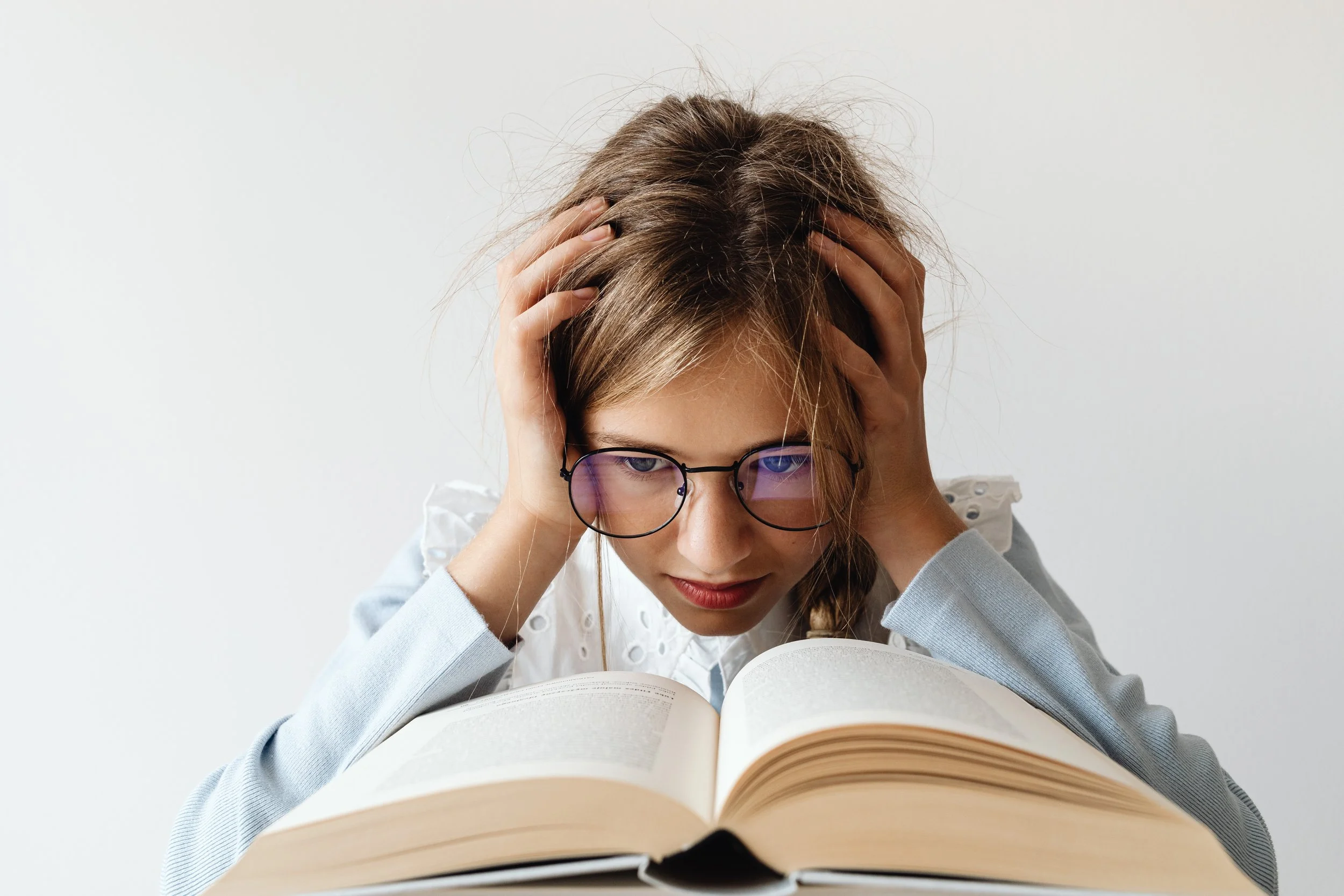 girl child wearing glasses with hands on her head in frustration while she reads a book in front of her