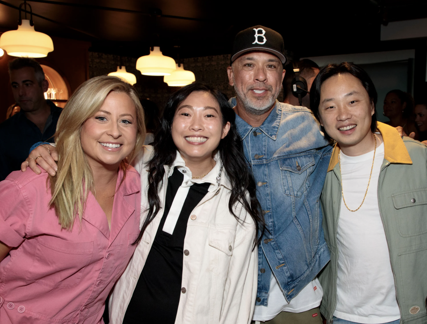 Four smiling people standing closely together inside a dimly lit room with hanging lights, with blurred background figures.