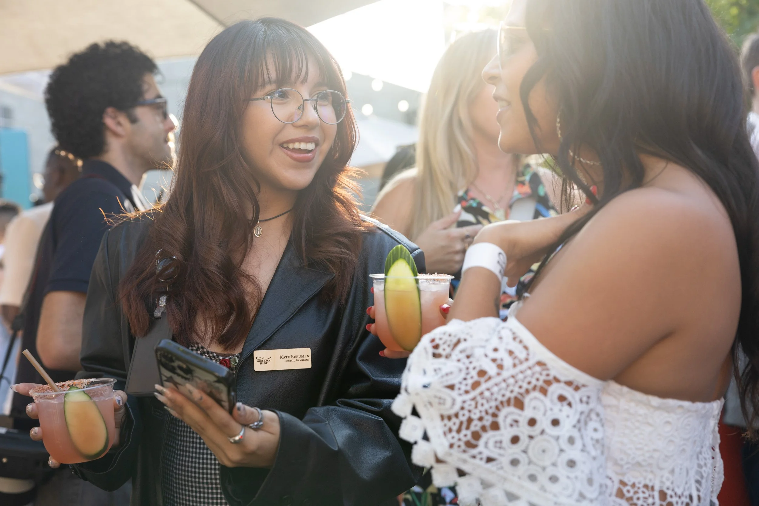 Two women are talking and holding drinks at an outdoor event. One woman has shoulder-length brown hair, glasses, and is wearing a jacket with a name tag. The other woman has long dark hair and is wearing a white lacy off-the-shoulder top. The woman with glasses is smiling and holding a smartphone.