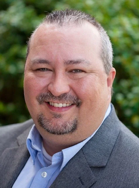 Headshot of a smiling man with short gray hair, a beard, and mustache, wearing a gray suit jacket and light blue shirt, outdoors with green foliage in the background.
