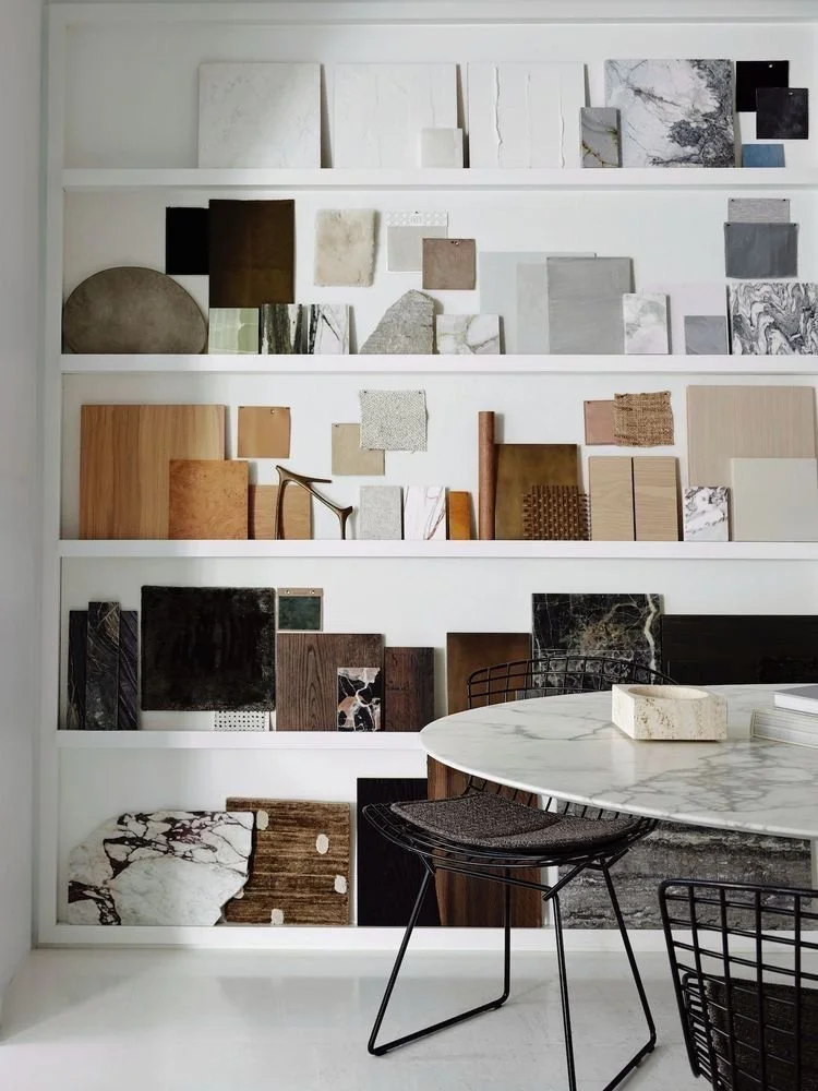 Étagère blanche avec divers échantillons de matériaux et de surfaces de différentes couleurs et textures, notamment marbre, bois, pierre, et métal, dans une pièce illuminée comportant également une table ronde en verre et des chaises en cuir noir.