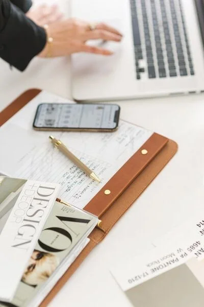 Main d'une femme utilisant un ordinateur portable, un smartphone à côté, ainsi qu'un carnet en cuir avec un stylo dessus, des magazines, et des papiers sur un bureau blanc.